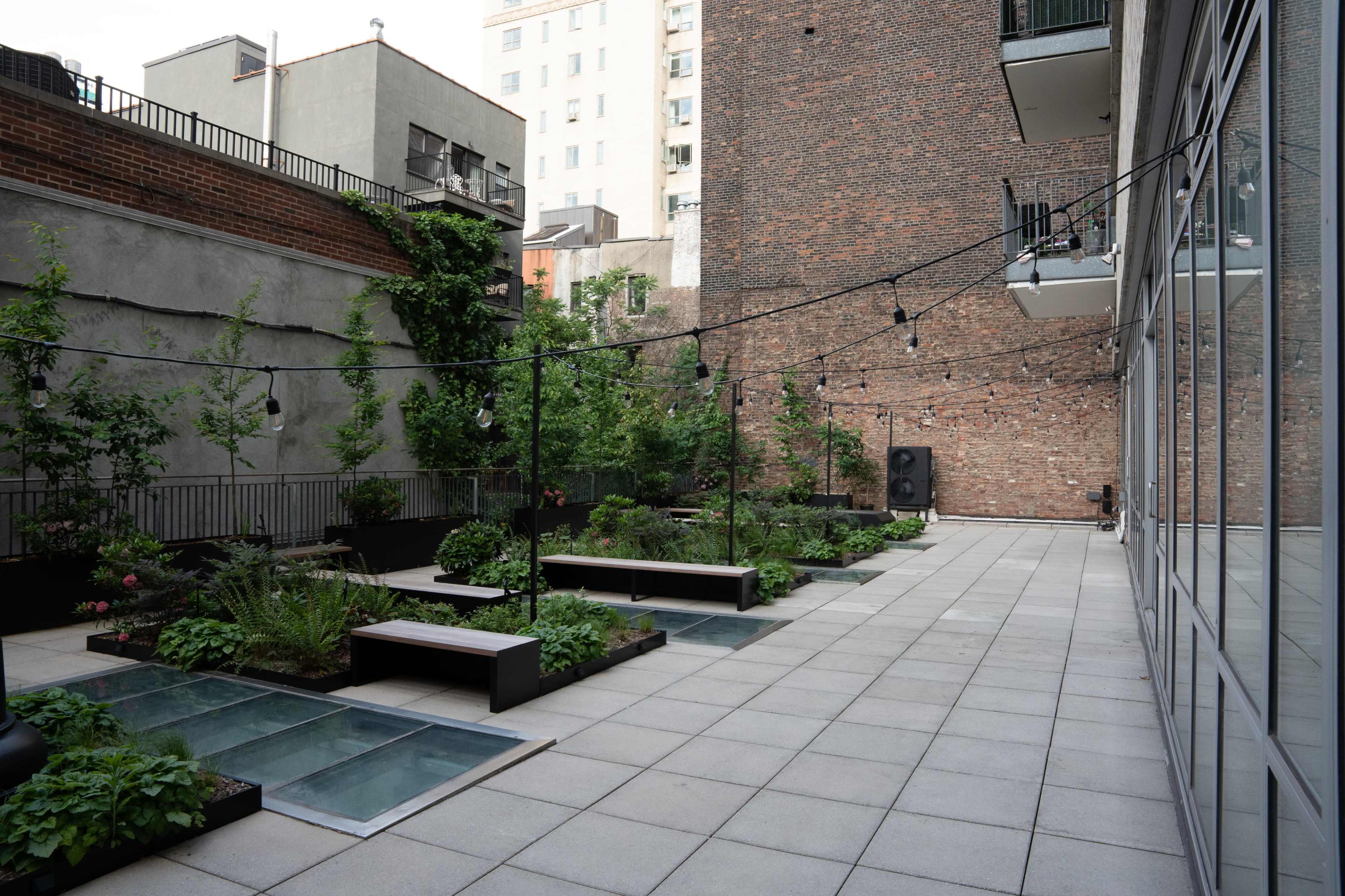 The image shows a rooftop garden with raised plant beds, benches, and string lights, surrounded by brick buildings.
