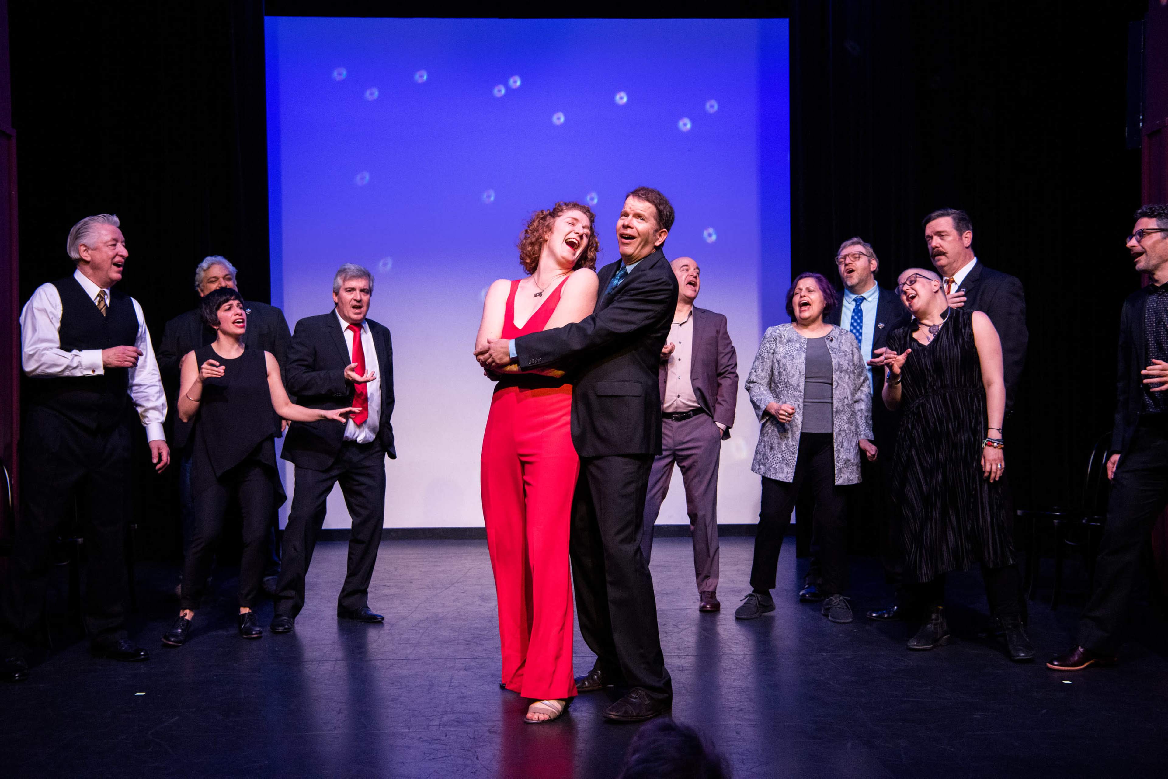 A group of performers stands on stage, with two in the foreground joyfully singing and dancing, while others look on in the background.
