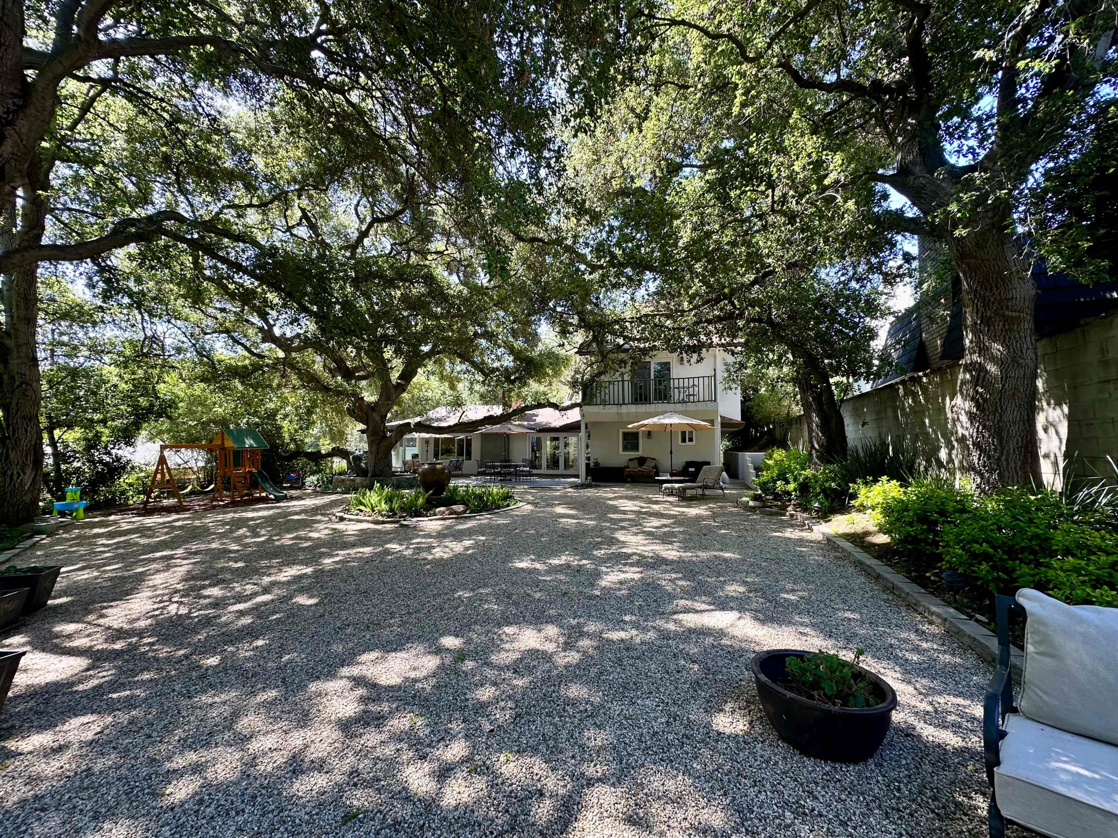 A spacious backyard features a gravel area with seating, surrounded by large trees and a two-story house in the background.