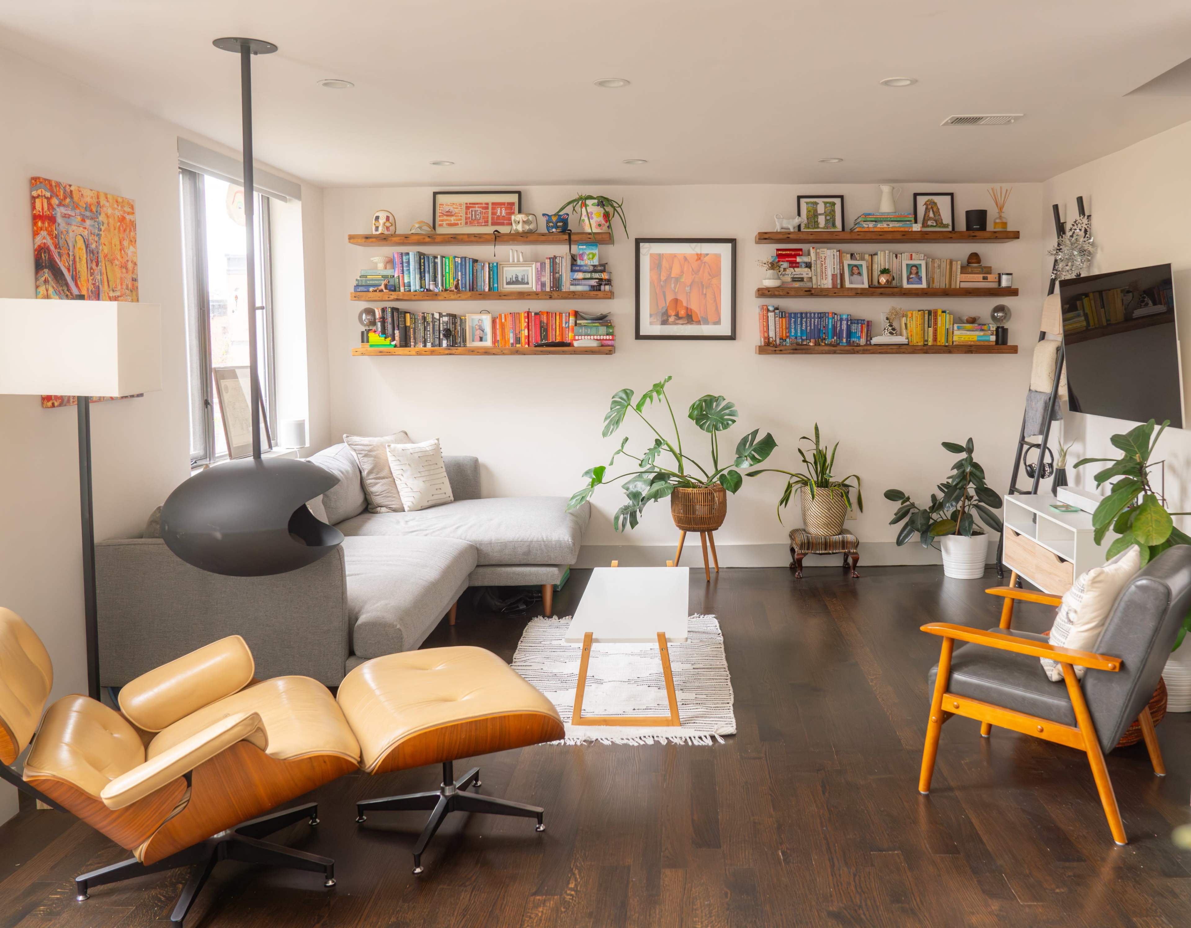 The image shows a modern living room with a gray sectional sofa, wooden shelves filled with books, plants, and a television mounted on the wall.