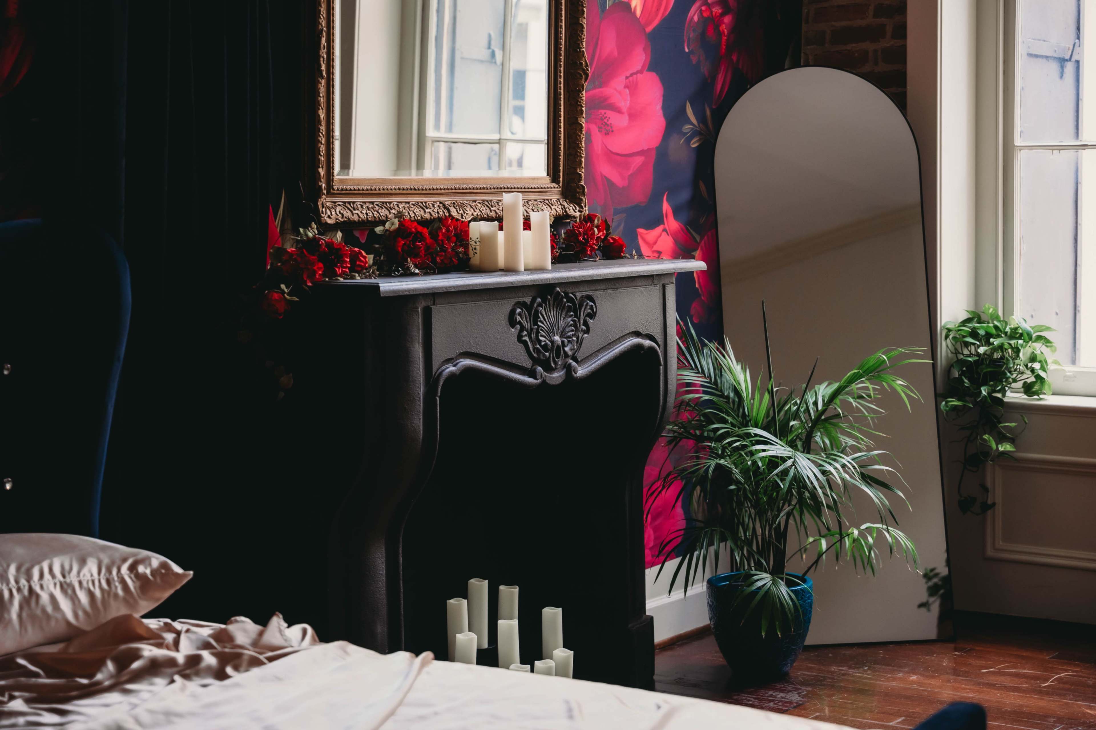 A dark fireplace with decorative candles and a mirror stands next to a bed in a room adorned with floral wallpaper.