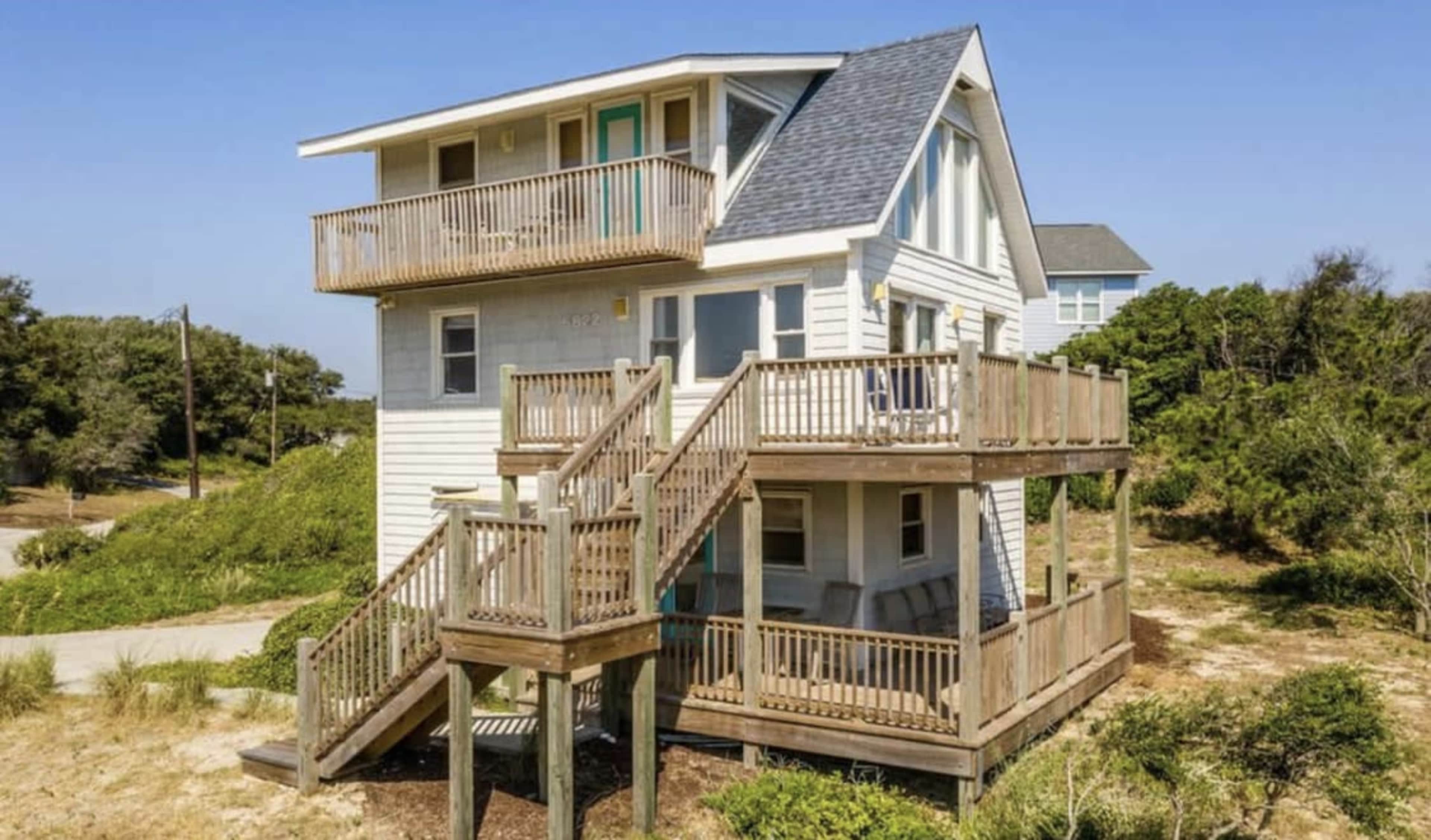 A two-story beach house with multiple balconies and wooden decks is situated among sandy vegetation.