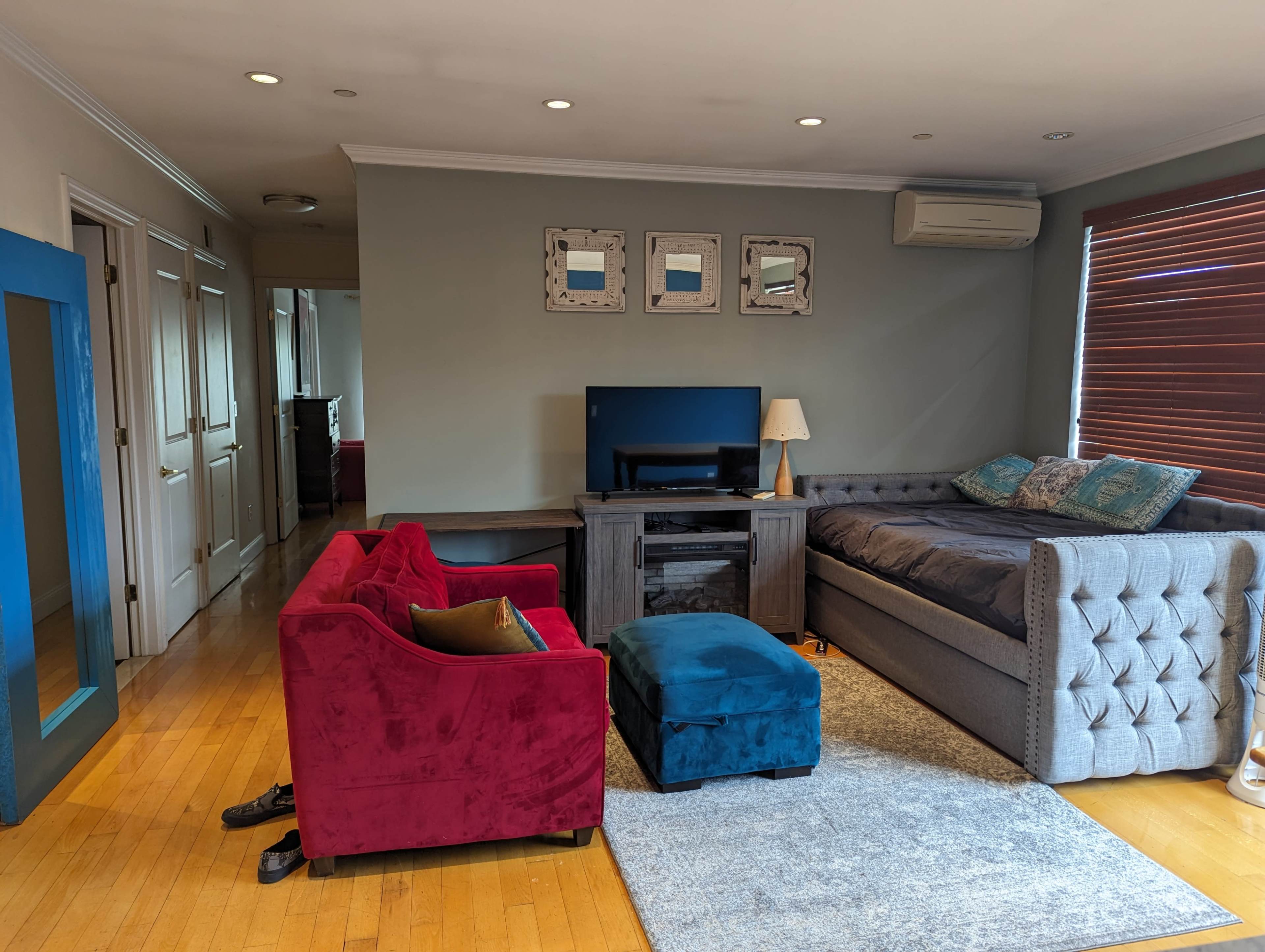 The living room features a gray sofa, a red chair, a blue ottoman, a wooden coffee table, and a television on a stand, with hardwood floors and wall art.