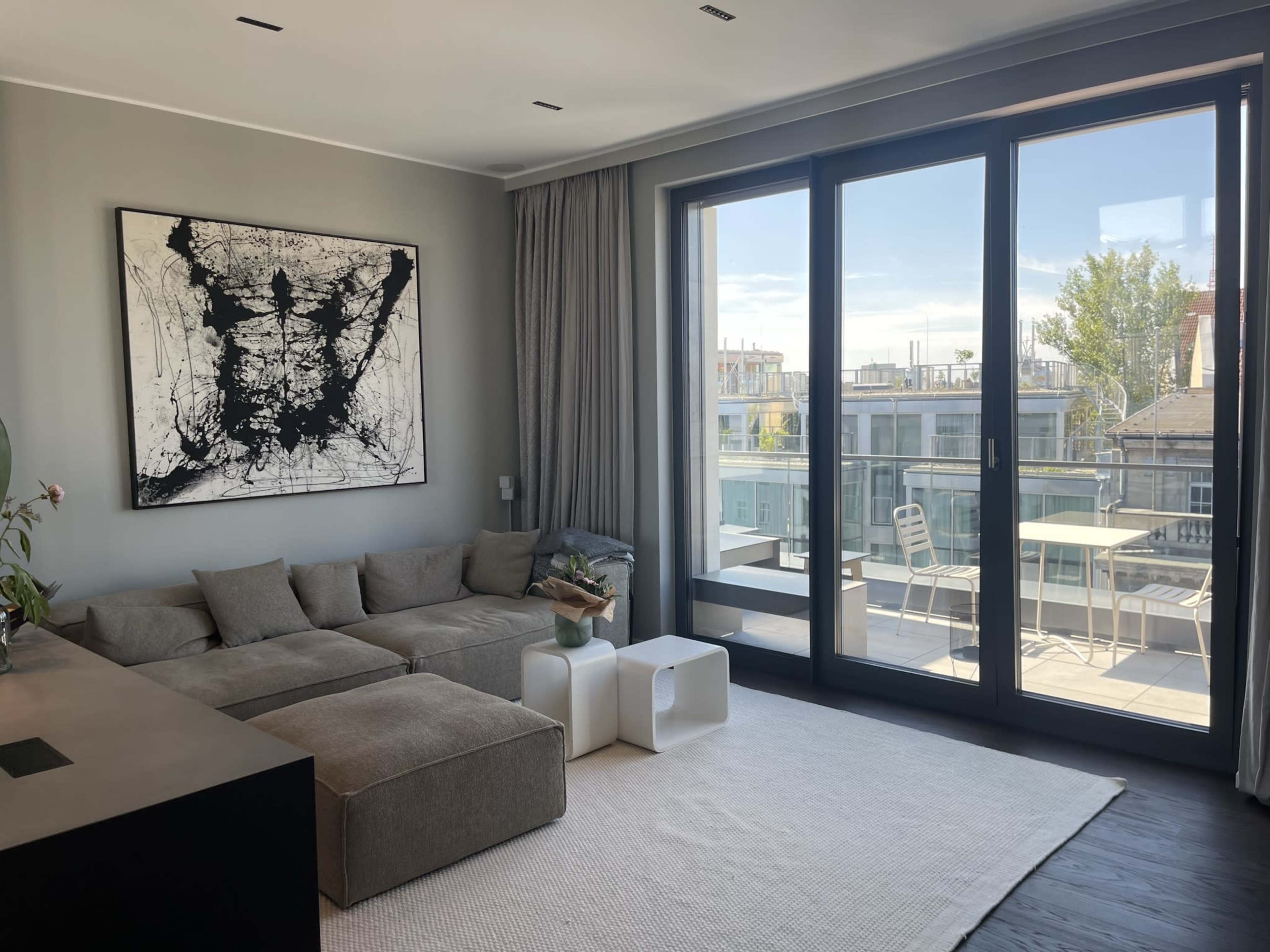 A modern living room features a large sectional sofa, a minimalist coffee table, and a large window that opens to a balcony with outdoor seating.