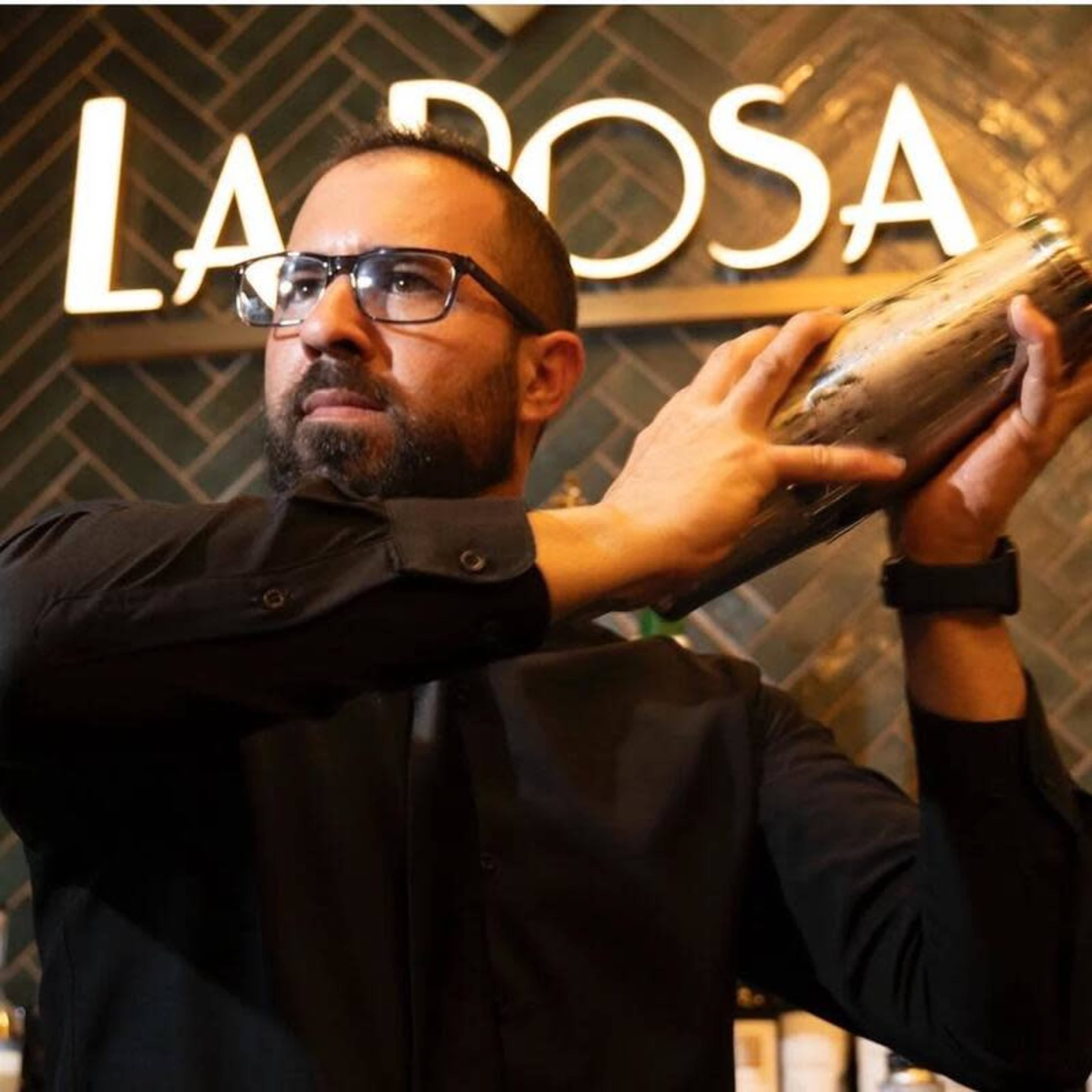 A bartender in a black shirt shakes a cocktail shaker in front of a sign that reads "LA ROSA."