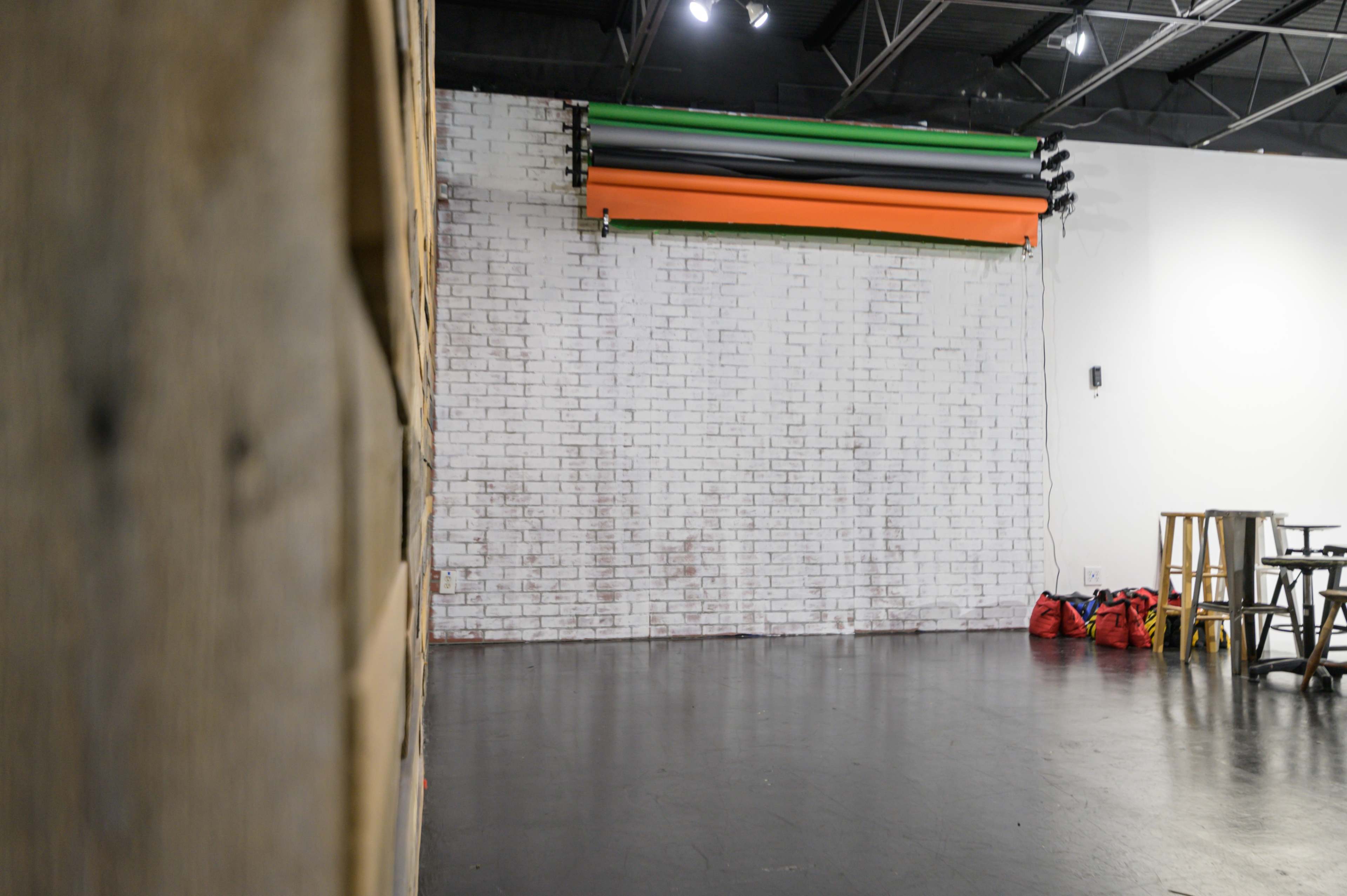 An empty studio space with a white brick wall, a roll of colorful backdrops, and red duffel bags in the corner.