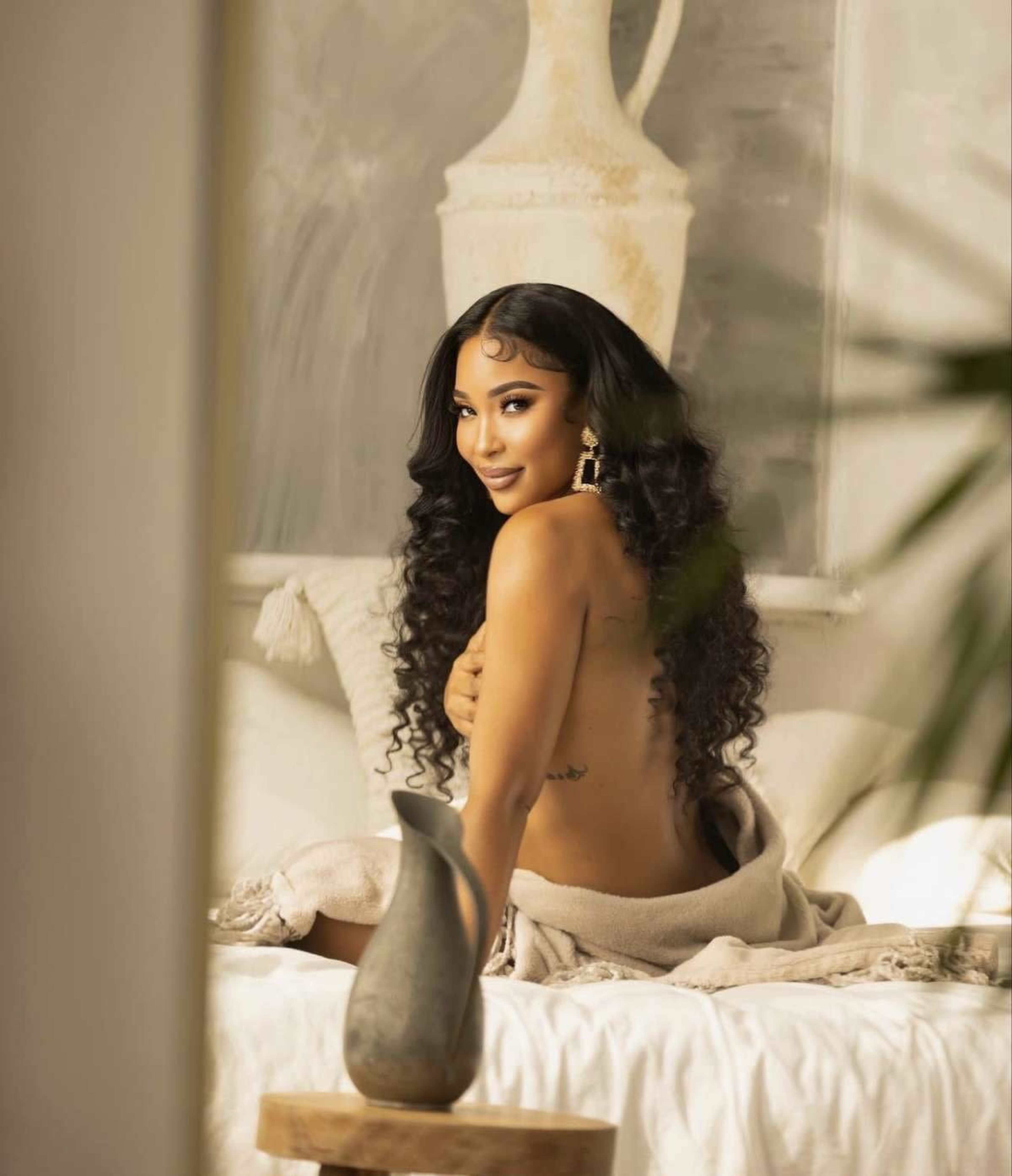 A person with long curly hair sits on a bed partially covered by a blanket, with decorative items and soft furnishings in the background.
