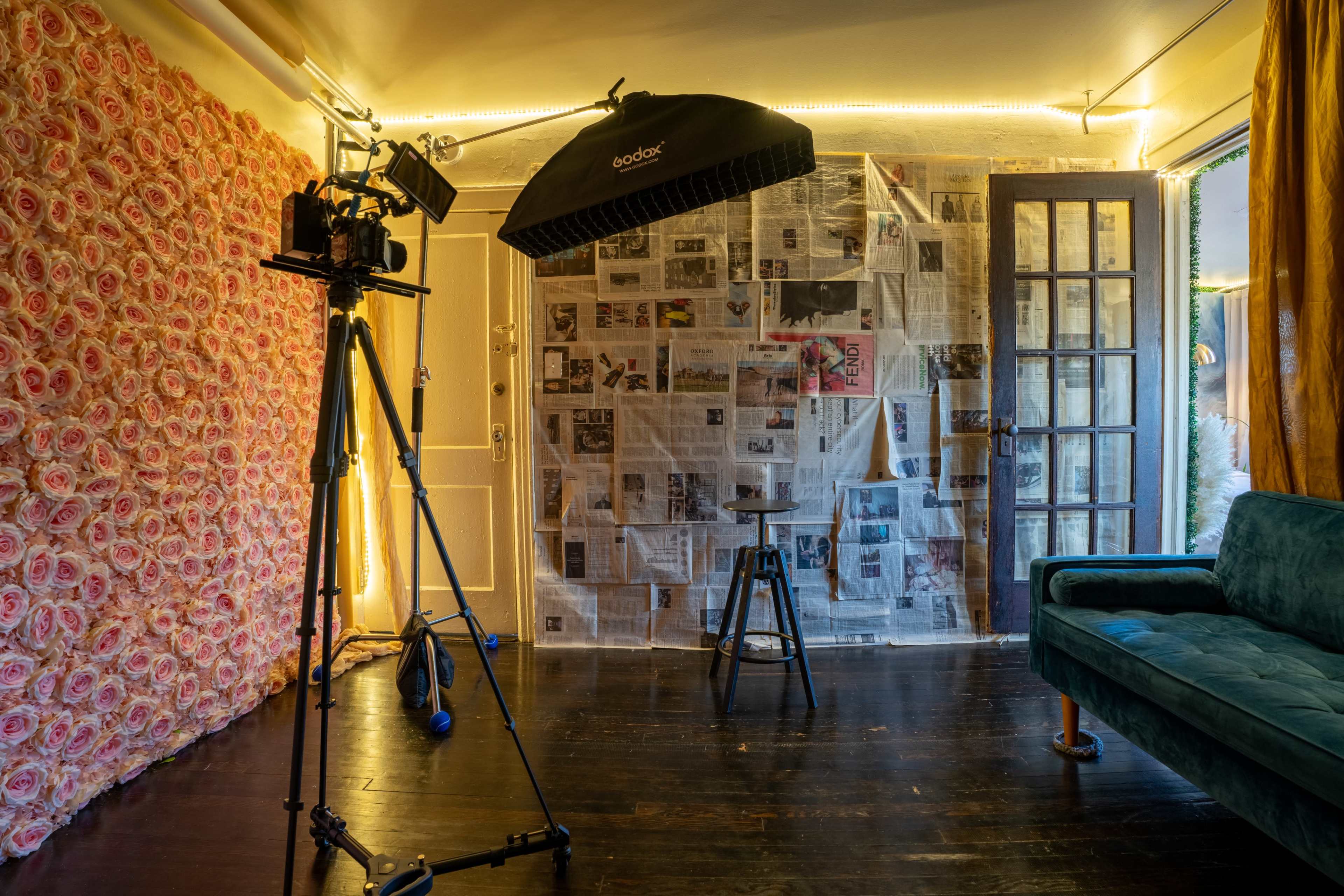 A photography studio with a camera on a tripod, a stool, and a backdrop made of newspapers, surrounded by a floral wall and illuminated by string lights.