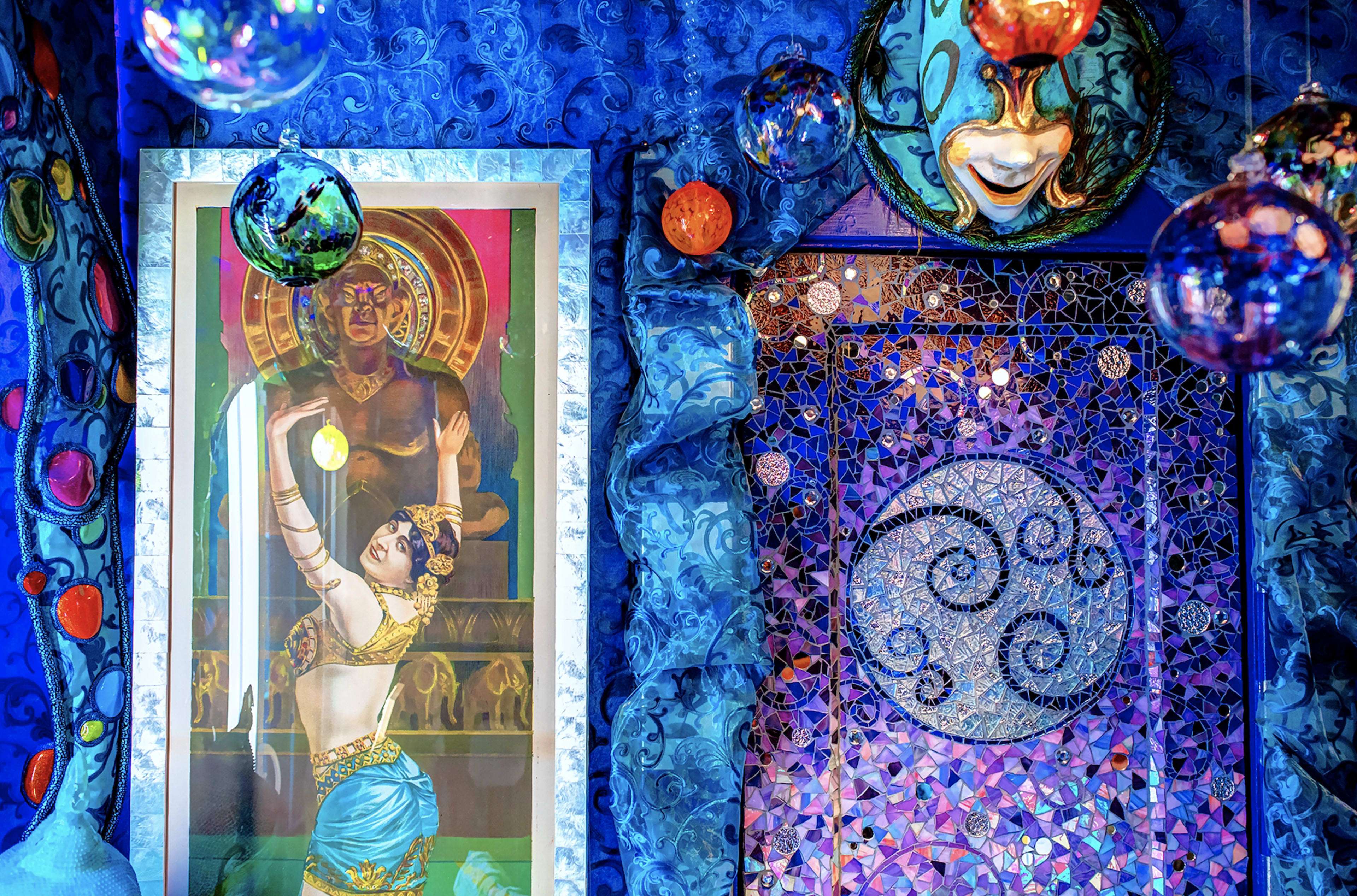 The scene features a vibrant blue interior with a framed artwork of a woman dancing next to a large depiction of a deity, surrounded by colorful orbs and decorative metalwork.