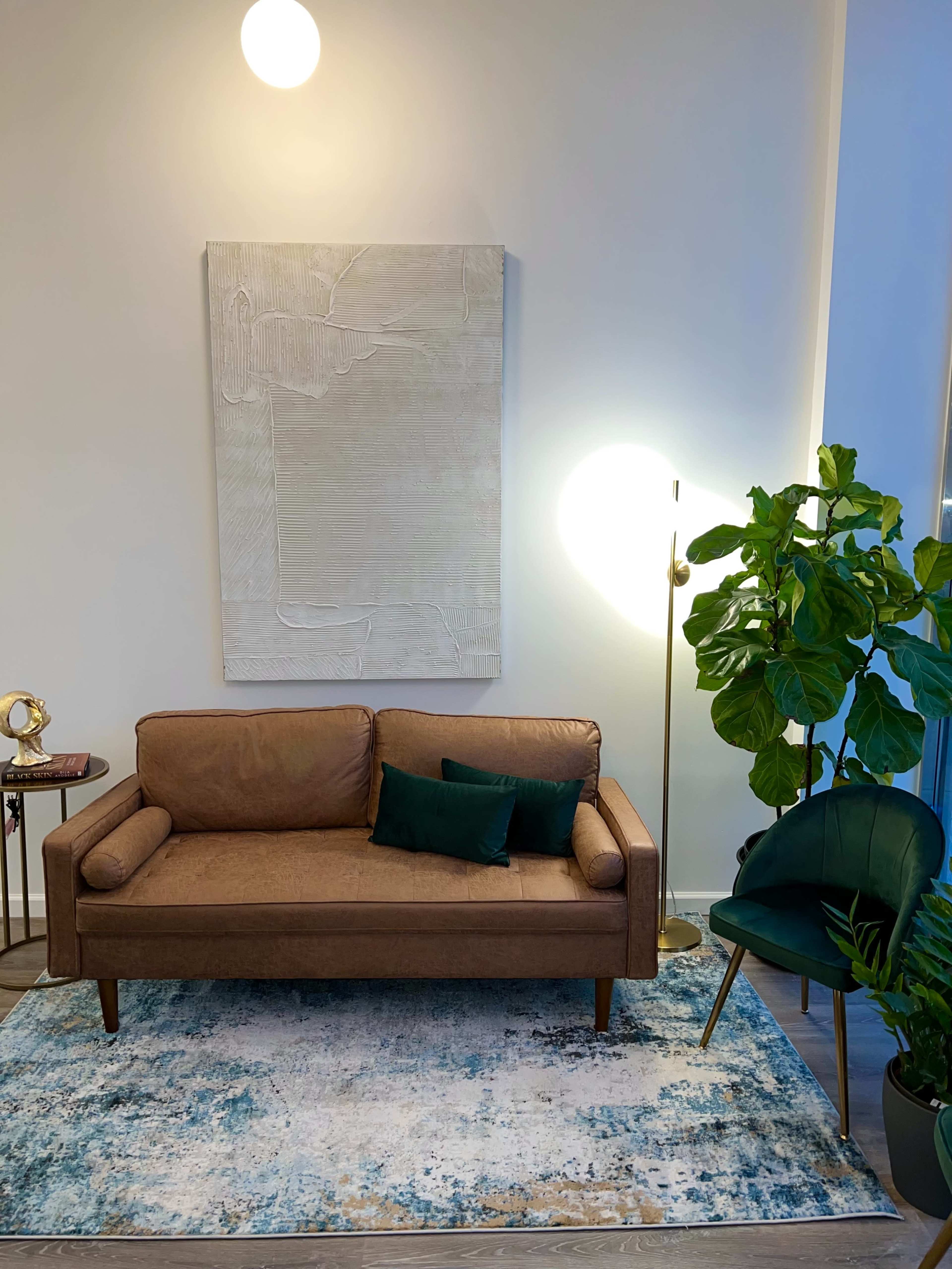 A brown sofa with a dark green cushion sits on a patterned rug next to a tall floor lamp and a large potted plant in a well-lit room.