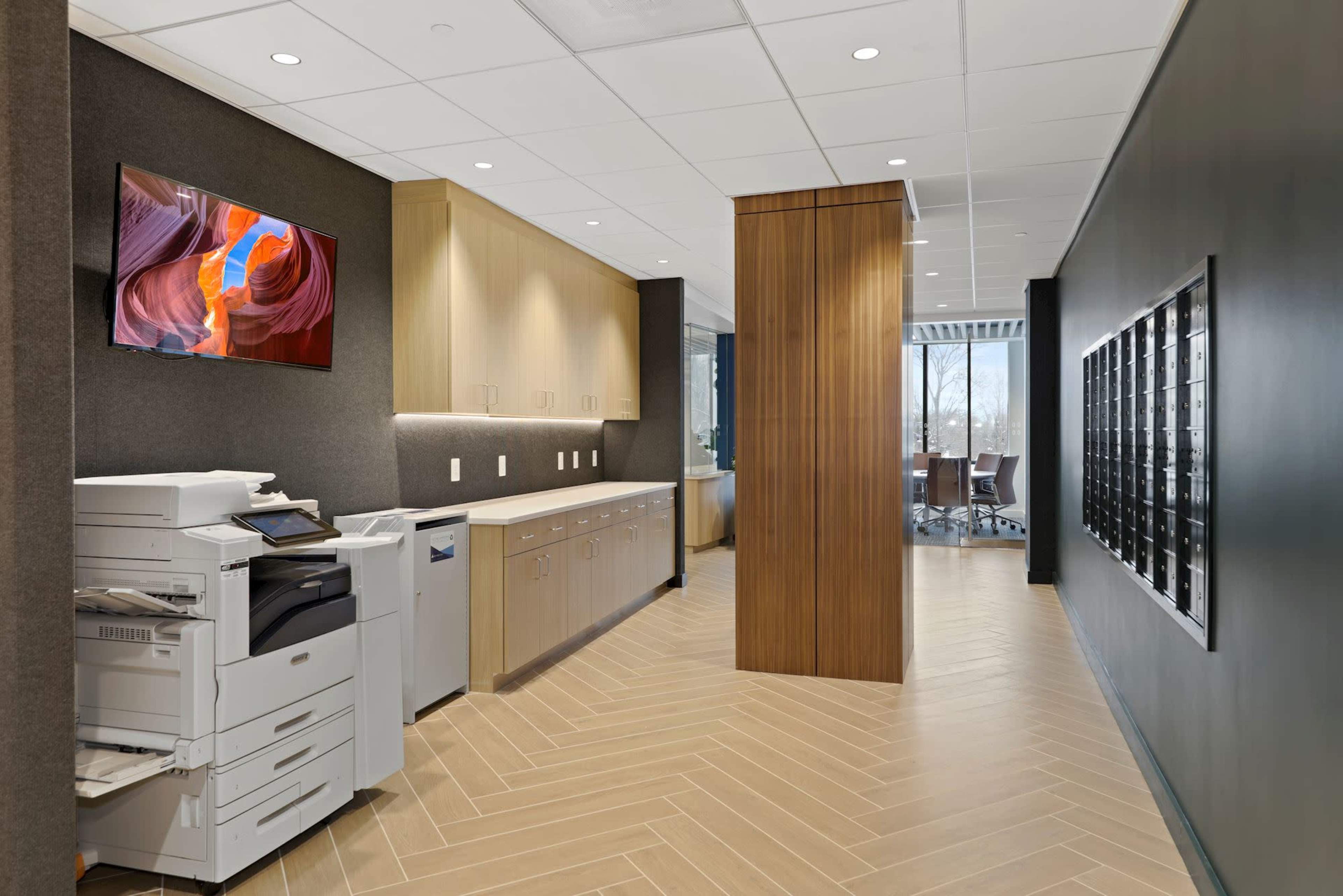 The image shows a modern office corridor featuring a printer station, wooden cabinets, and an adjacent meeting room.
