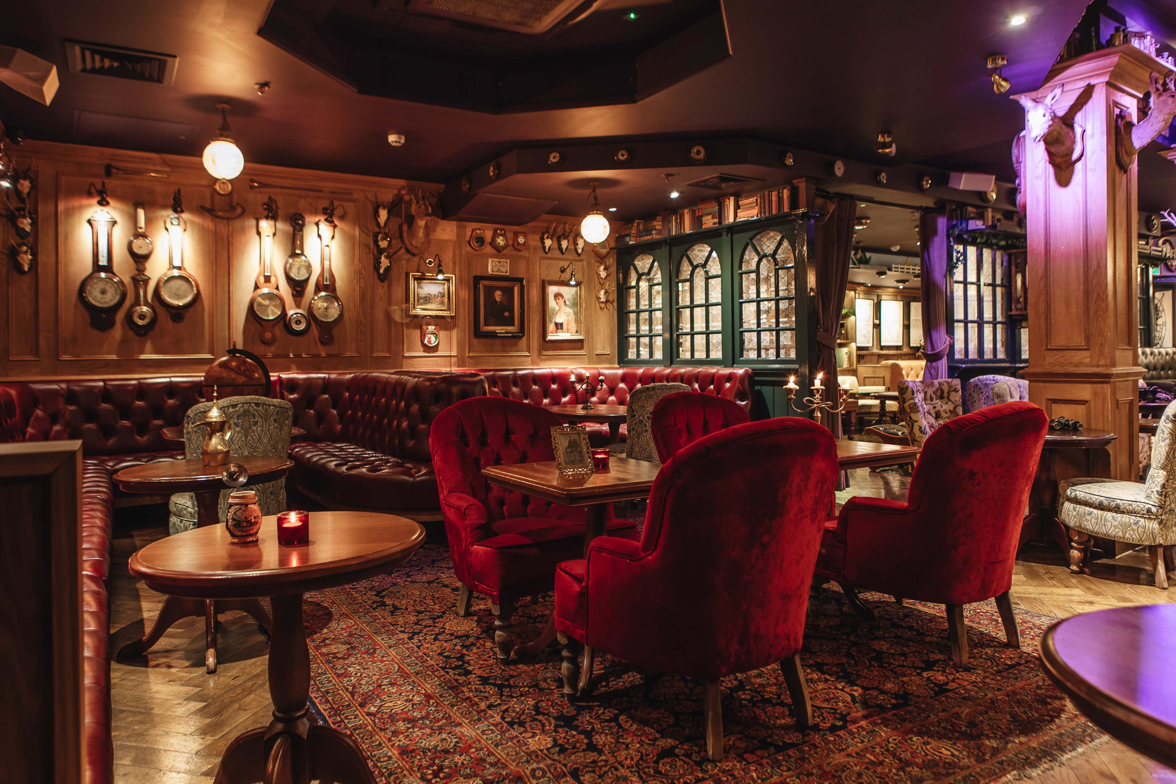 The image shows a cozy bar interior with plush red chairs, dark wood furnishings, and decorative wall hangings.