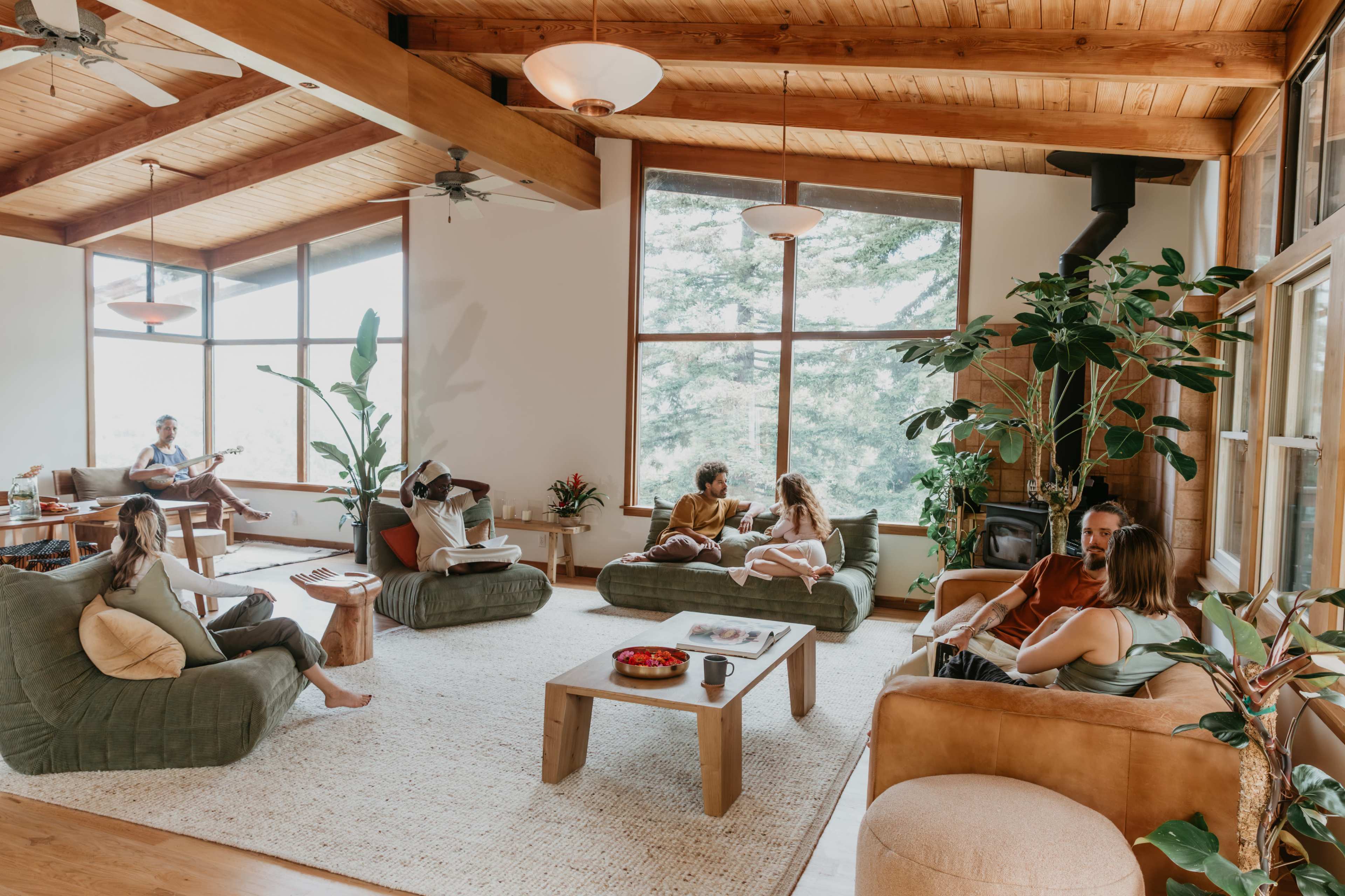 A spacious living room features a group of people engaging in conversation among large windows, plants, and comfortable seating.