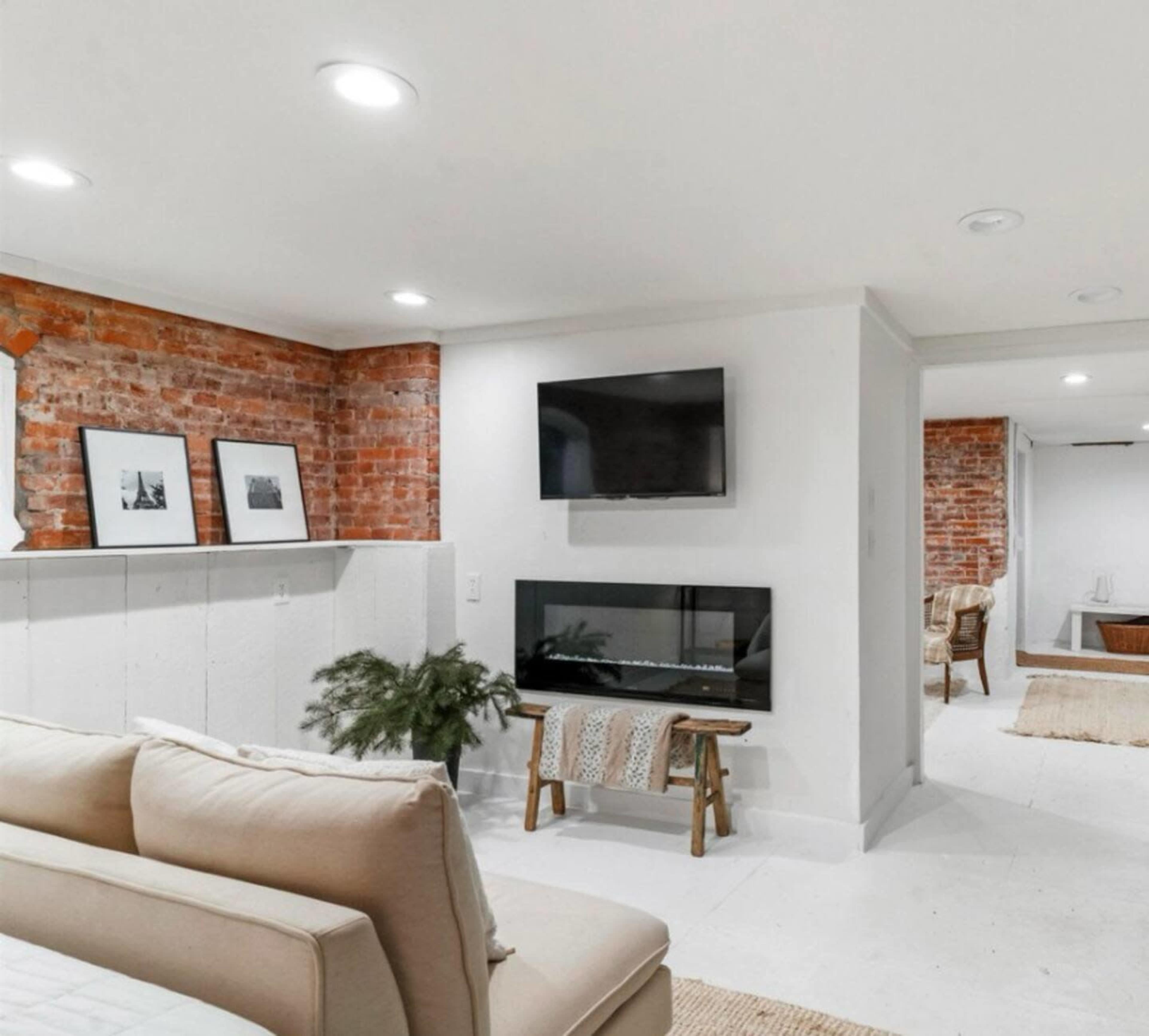 A modern basement area featuring exposed brick walls, a mounted television, a linear electric fireplace, and neutral-colored furnishings.