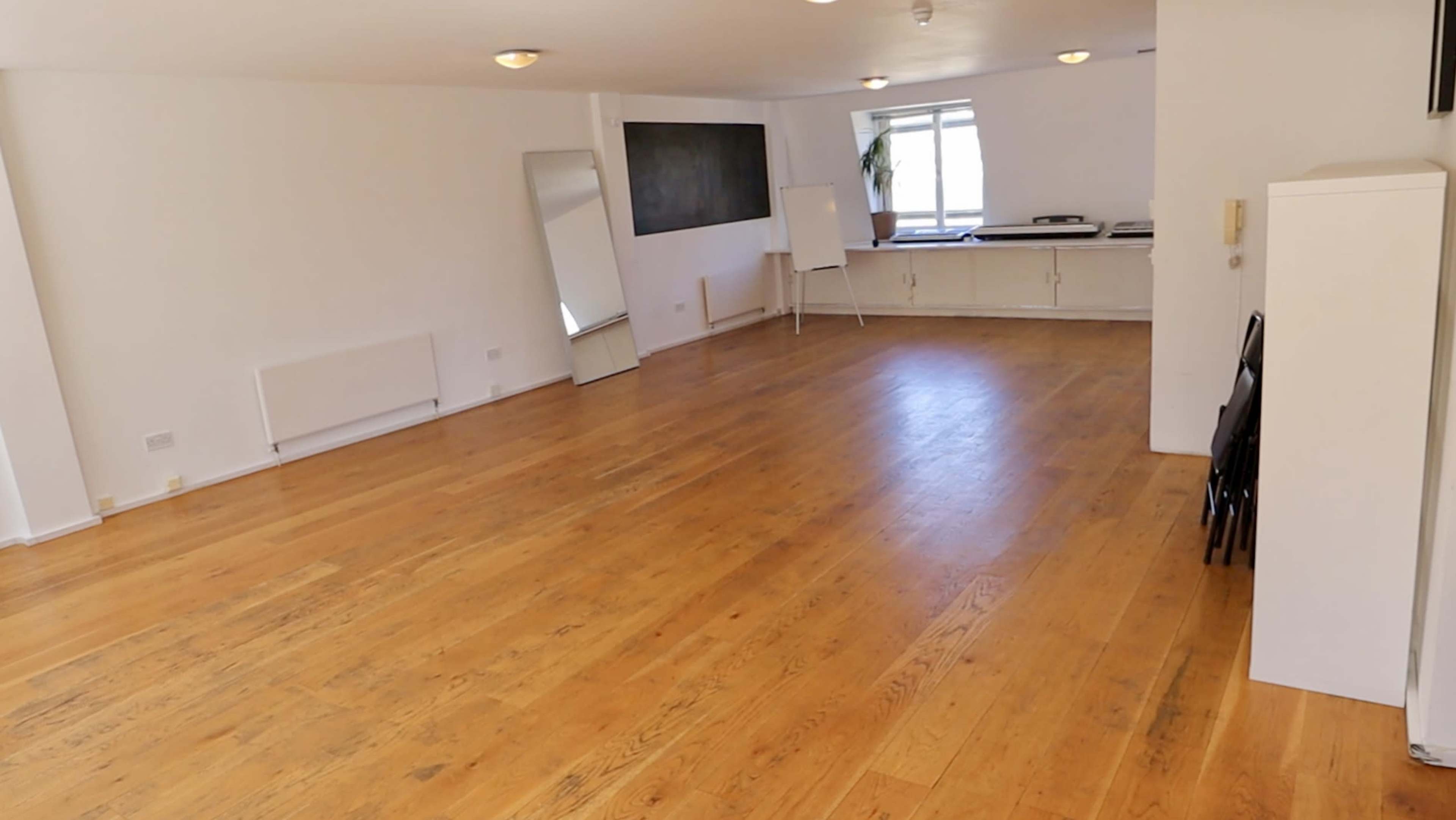A spacious, empty room with wooden flooring, large windows, a mirror, and a whiteboard at one end.