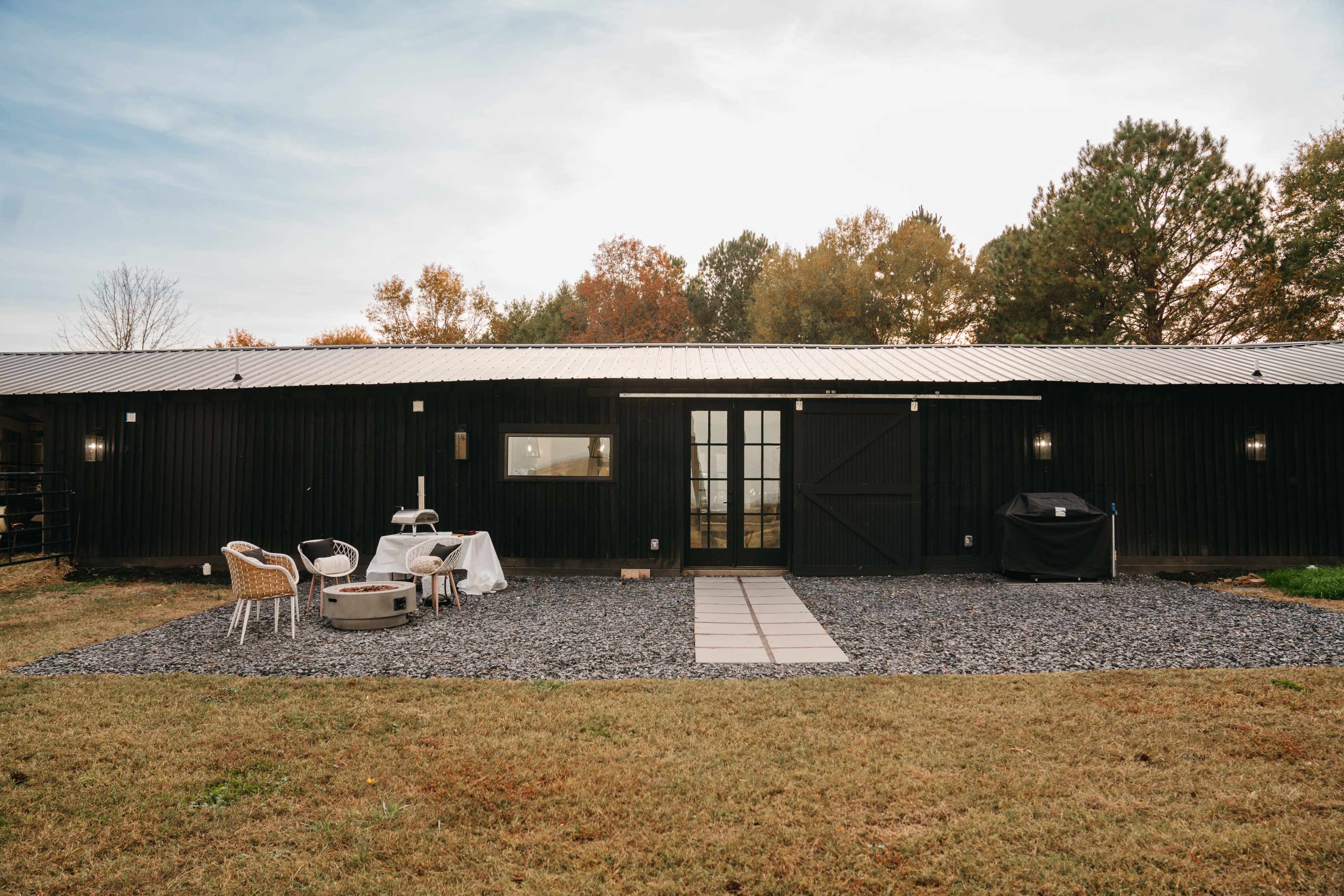 A black wooden cabin with a patio area featuring outdoor furniture and a barbecue grill, surrounded by gravel and grass.