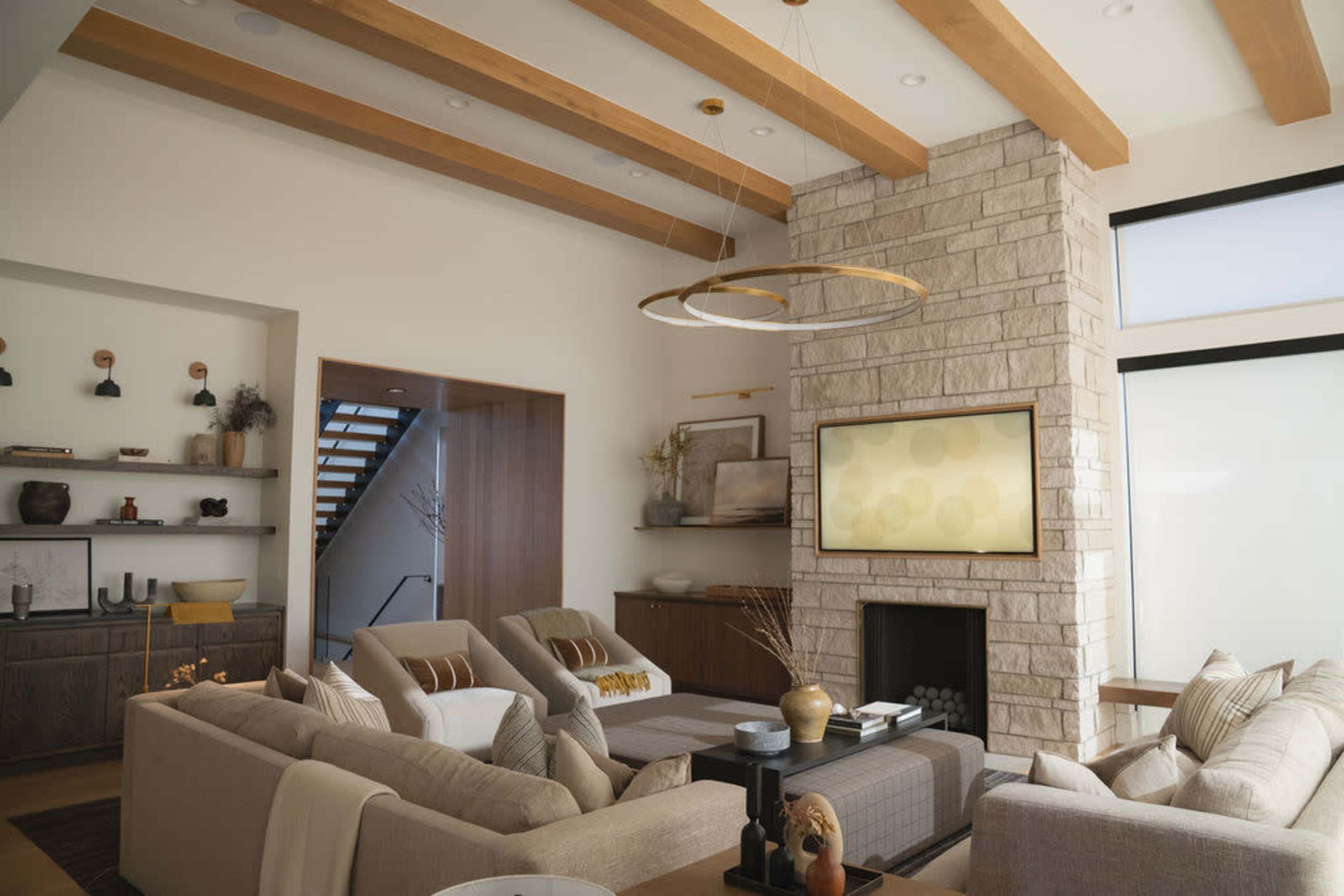 The image shows a modern living room with a stone fireplace, a flat-screen TV, beige sofas, and wooden ceiling beams.