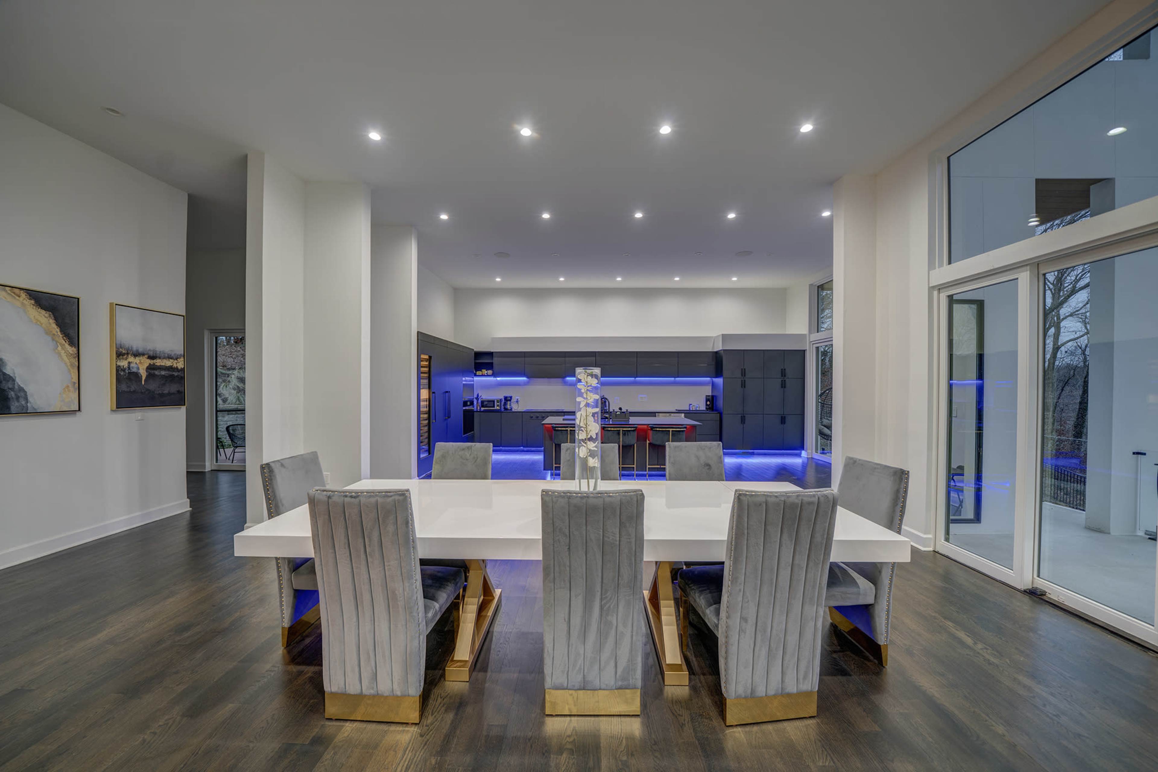 A modern dining area features a large white table surrounded by eight upholstered chairs, with a kitchen in the background illuminated by blue lighting.