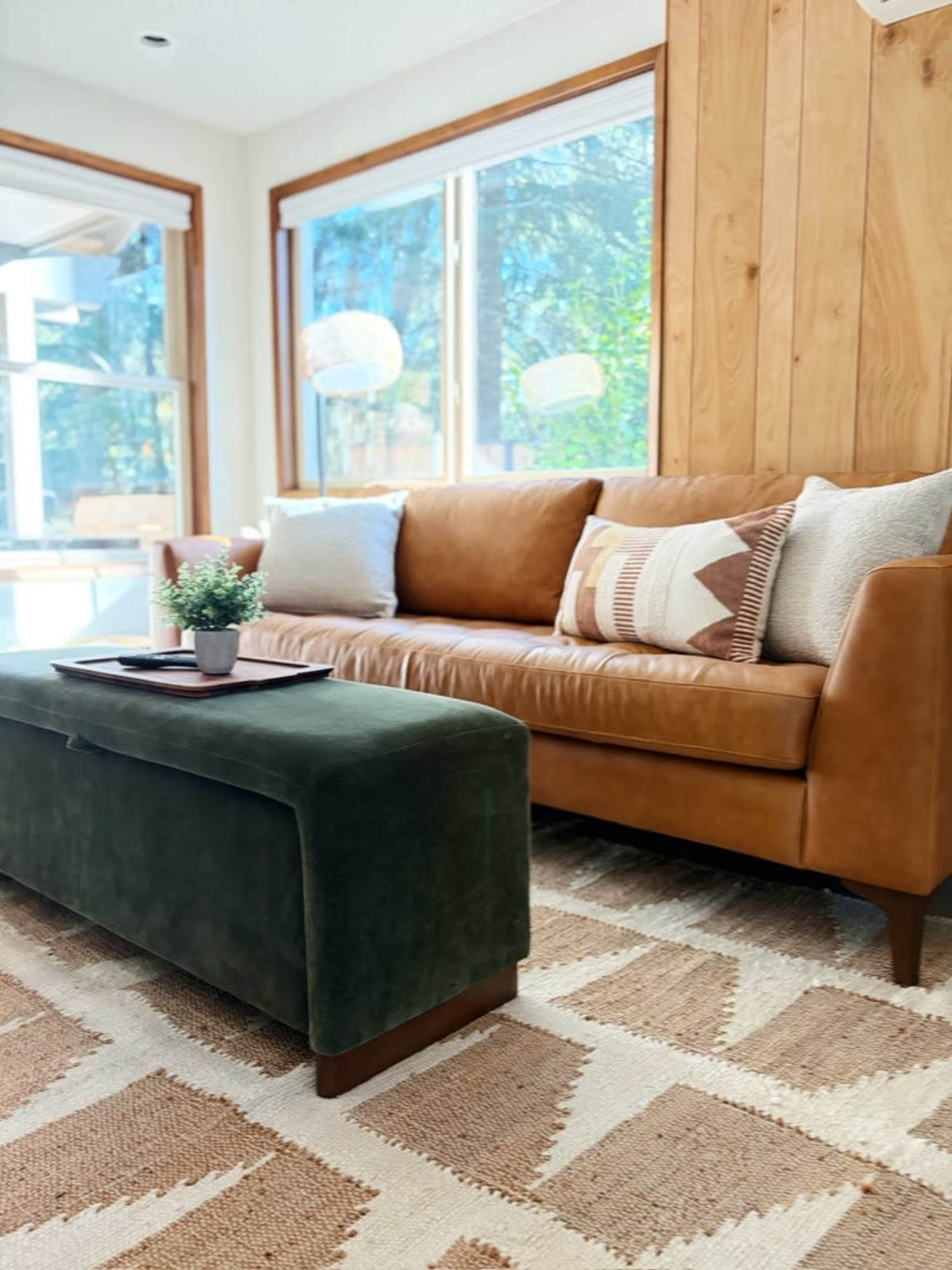 The image shows a living room with a brown leather couch, a green ottoman, and a small plant on a coffee table, surrounded by large windows.