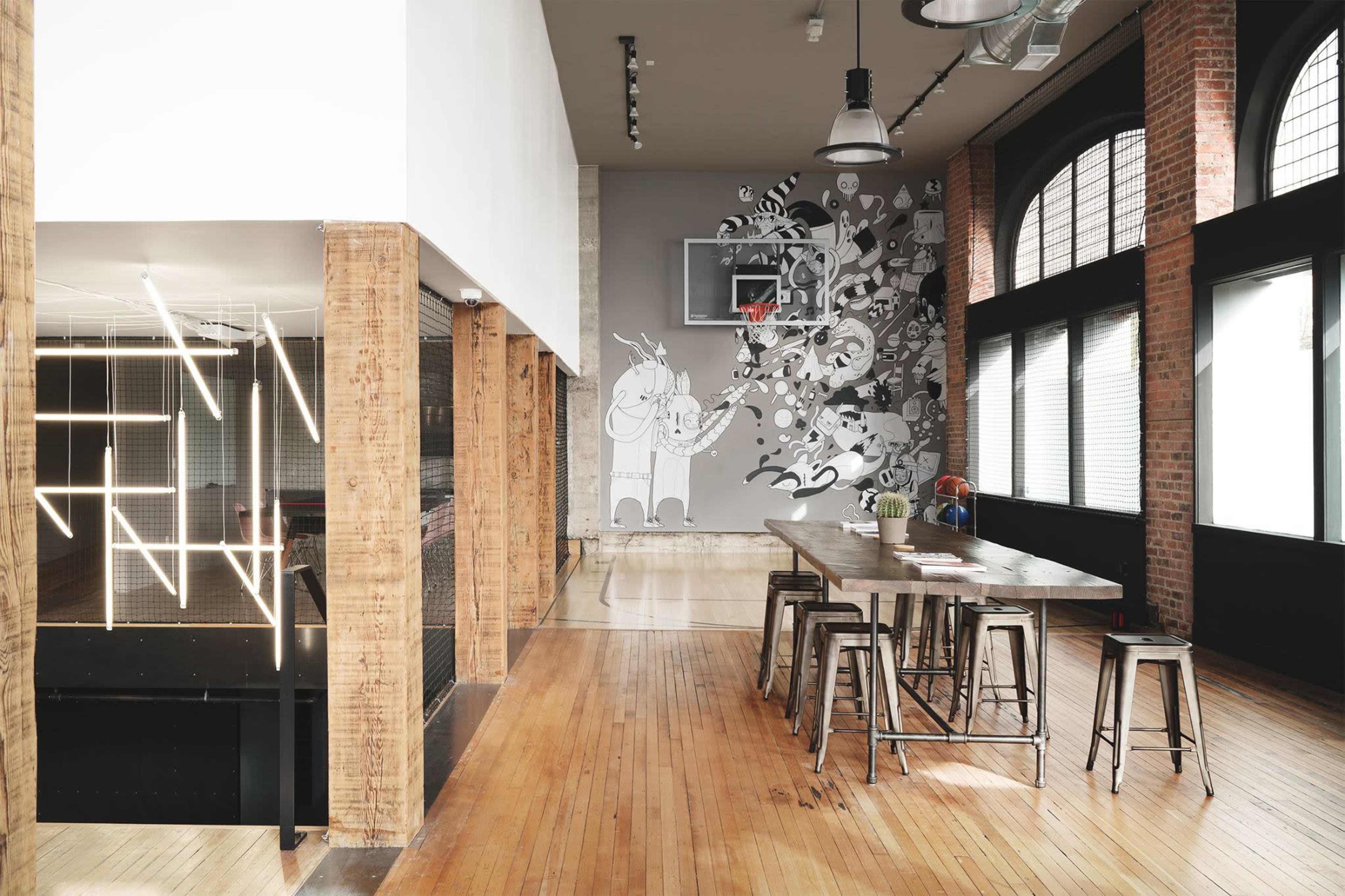 The image shows a spacious, modern interior featuring a long wooden table, metal stools, and a wall mural, with a basketball hoop in the corner and large windows allowing natural light.