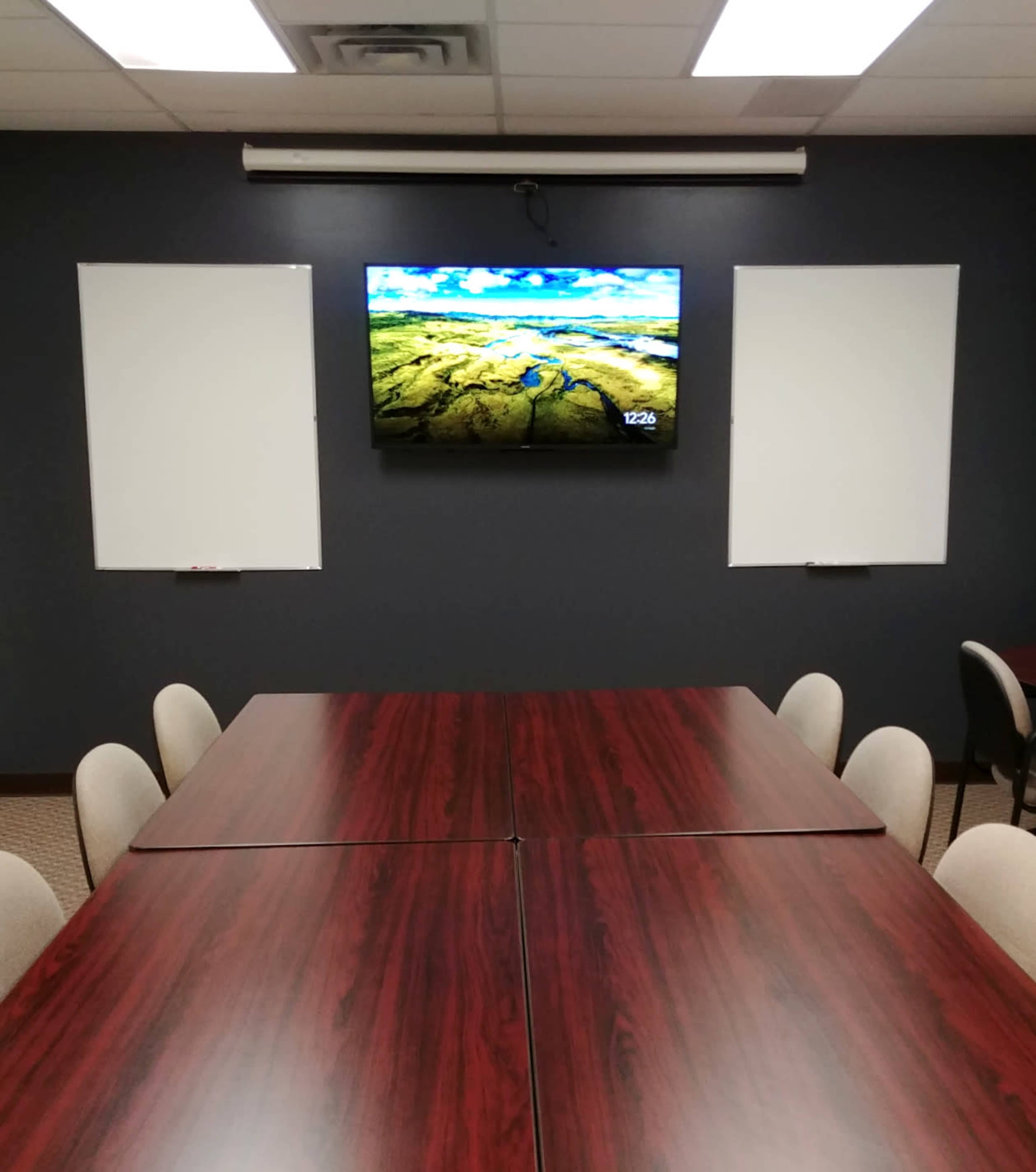 The image shows a conference room with two long tables arranged in a U-shape, a television mounted on the wall displaying a landscape, and two blank whiteboards on either side.