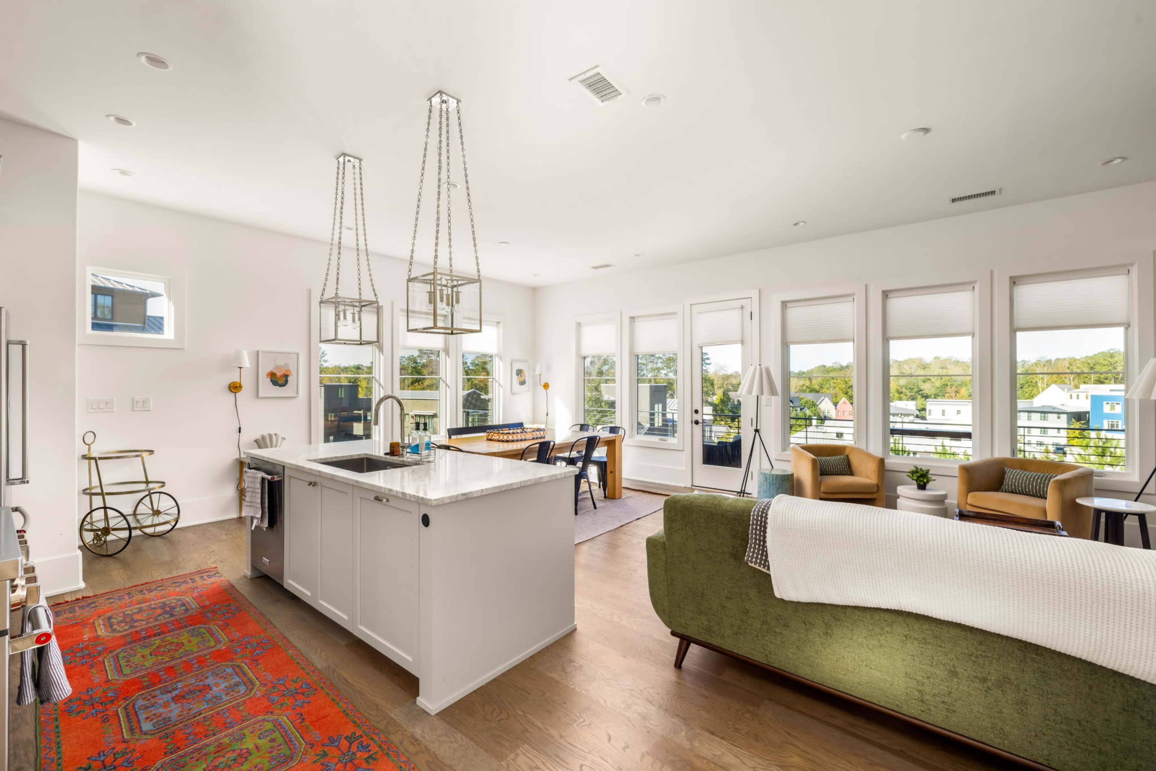 The image shows a bright and modern open-concept living space with a kitchen, dining area, and seating arrangement near large windows.