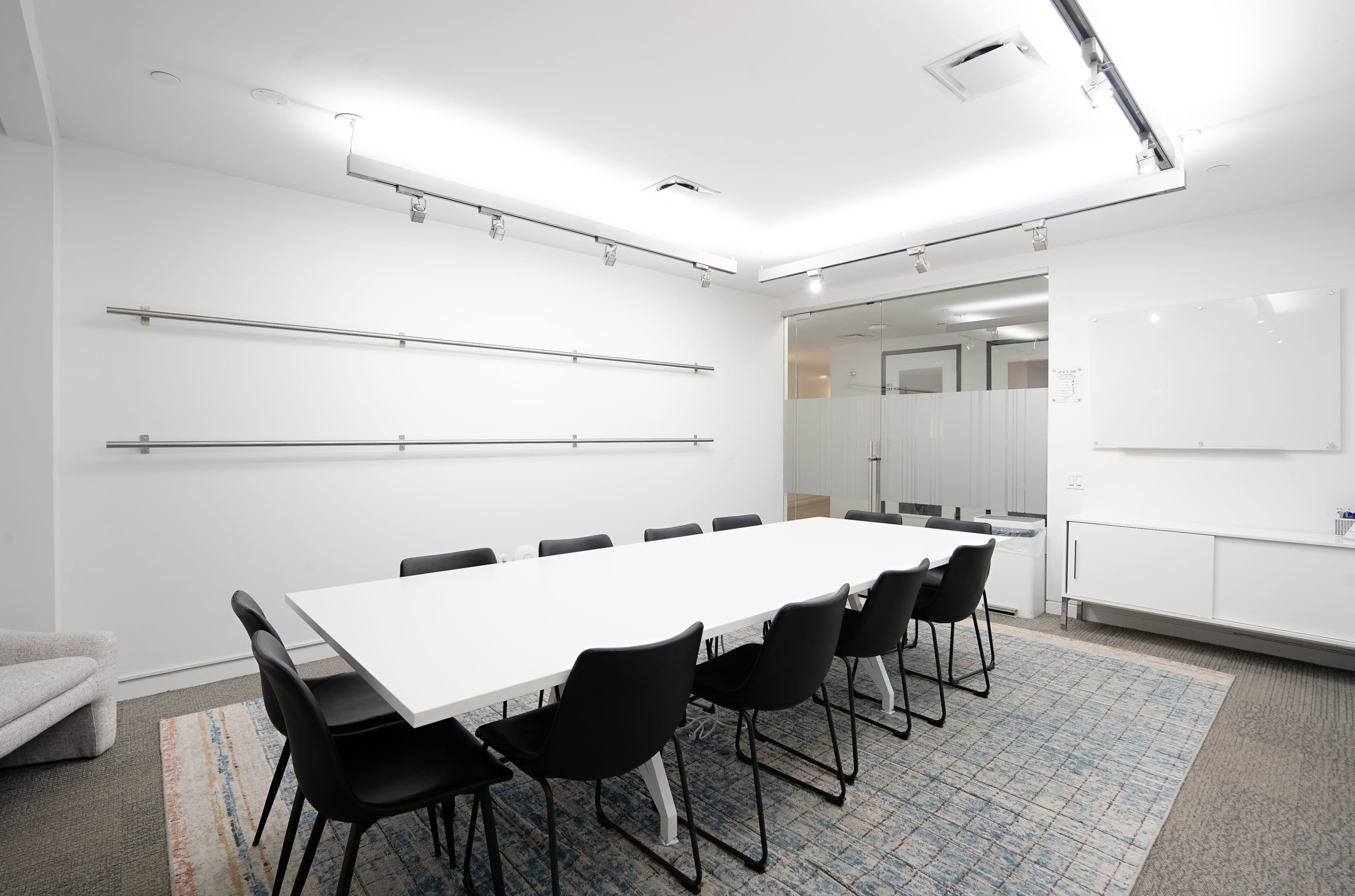 A modern conference room features a long white table surrounded by black chairs, with minimalist decor and a light-colored rug.