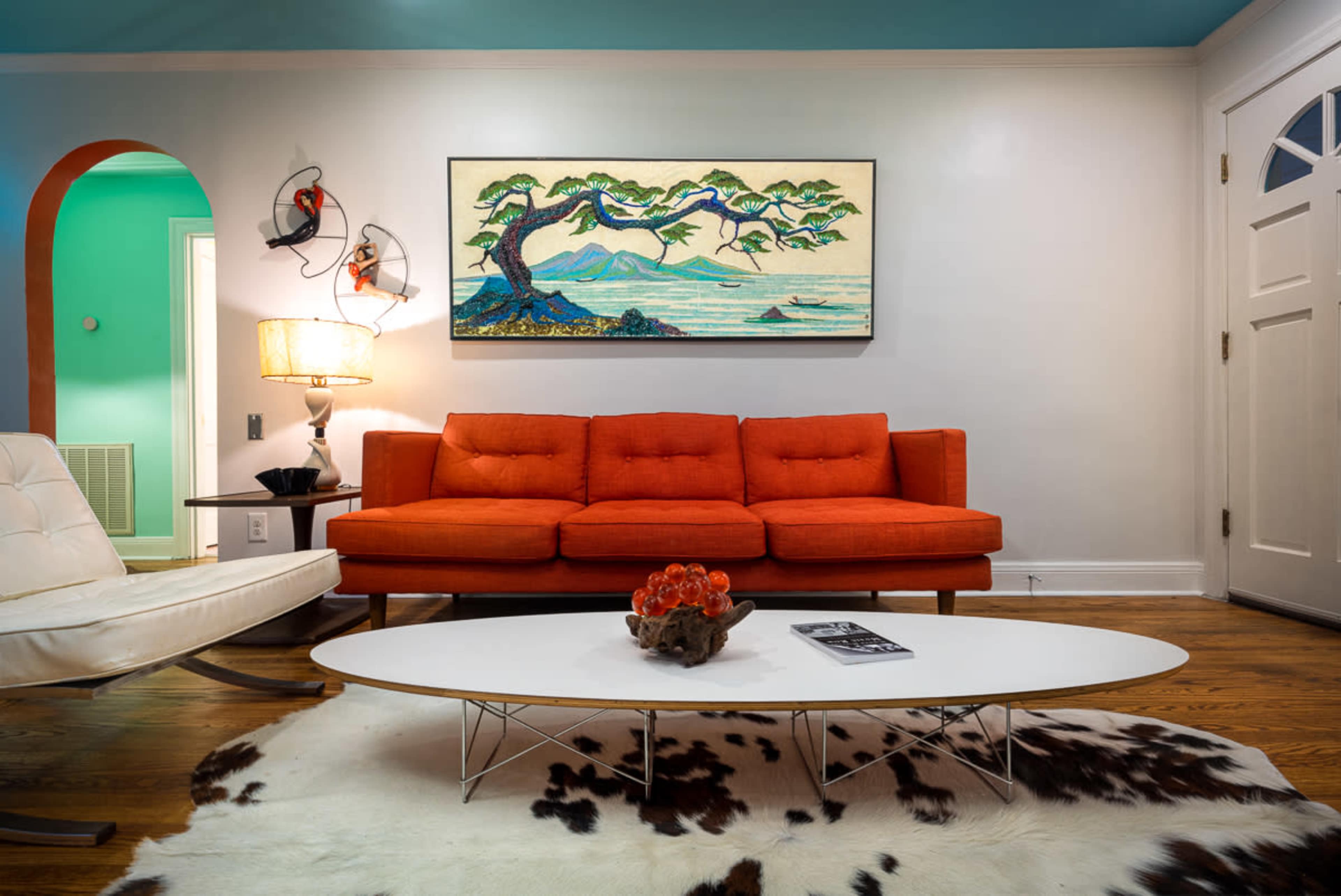 A modern living room featuring an orange sofa, a white coffee table, and a large artwork depicting a coastal scene on the wall.