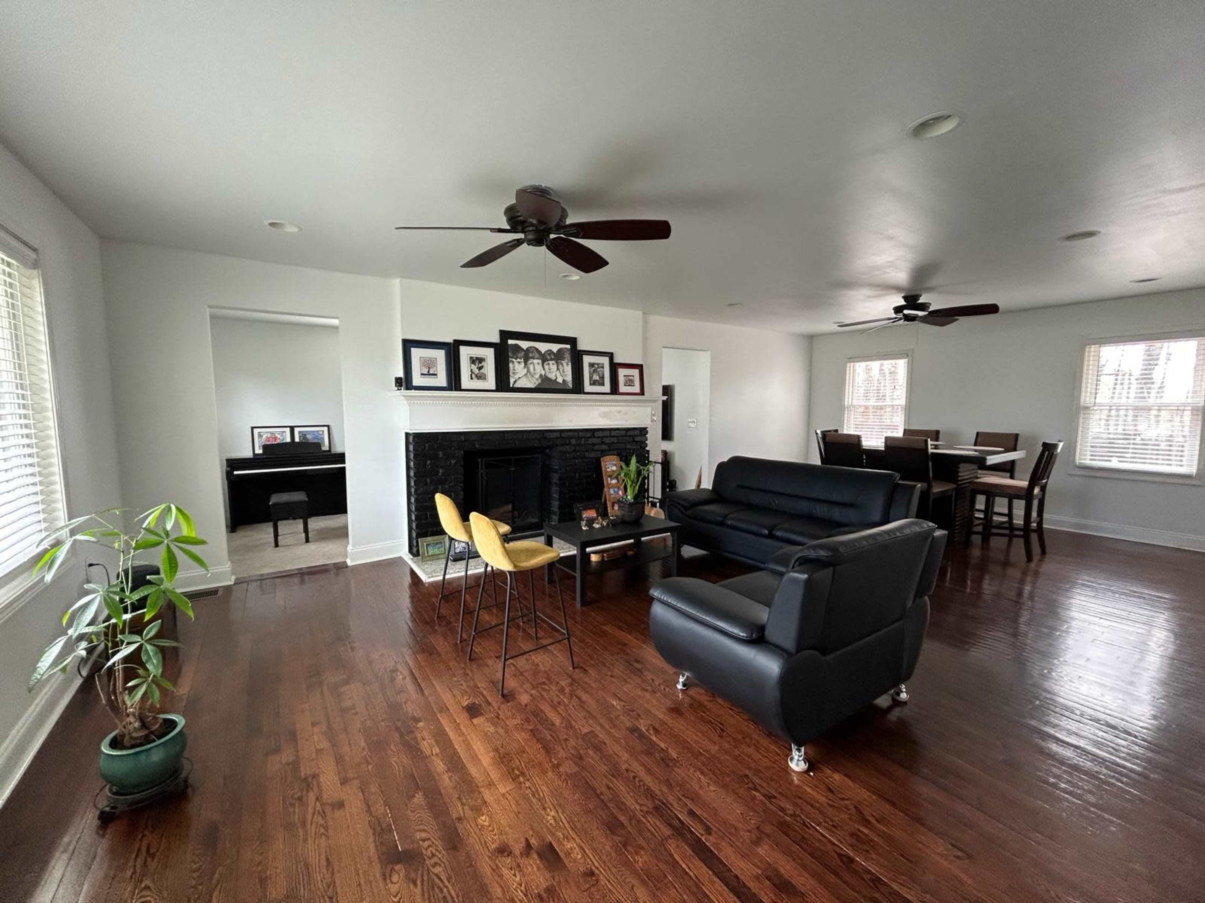 A spacious living room features dark wooden floors, a black fireplace with framed artwork above it, two ceiling fans, and modern furniture including a black couch and yellow chair.