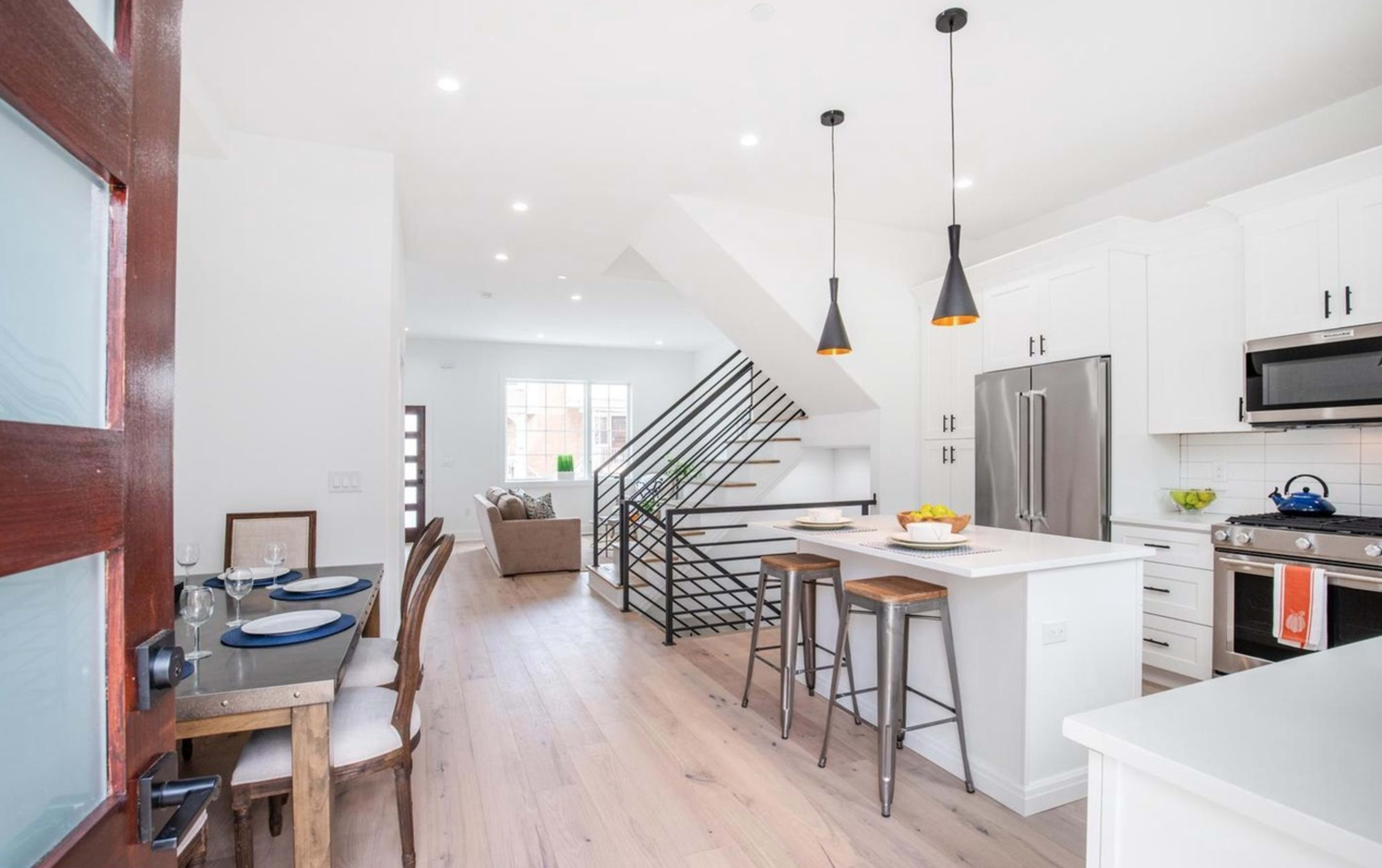 The image shows a modern kitchen with white cabinets, stainless steel appliances, and a dining area adjacent to a staircase.