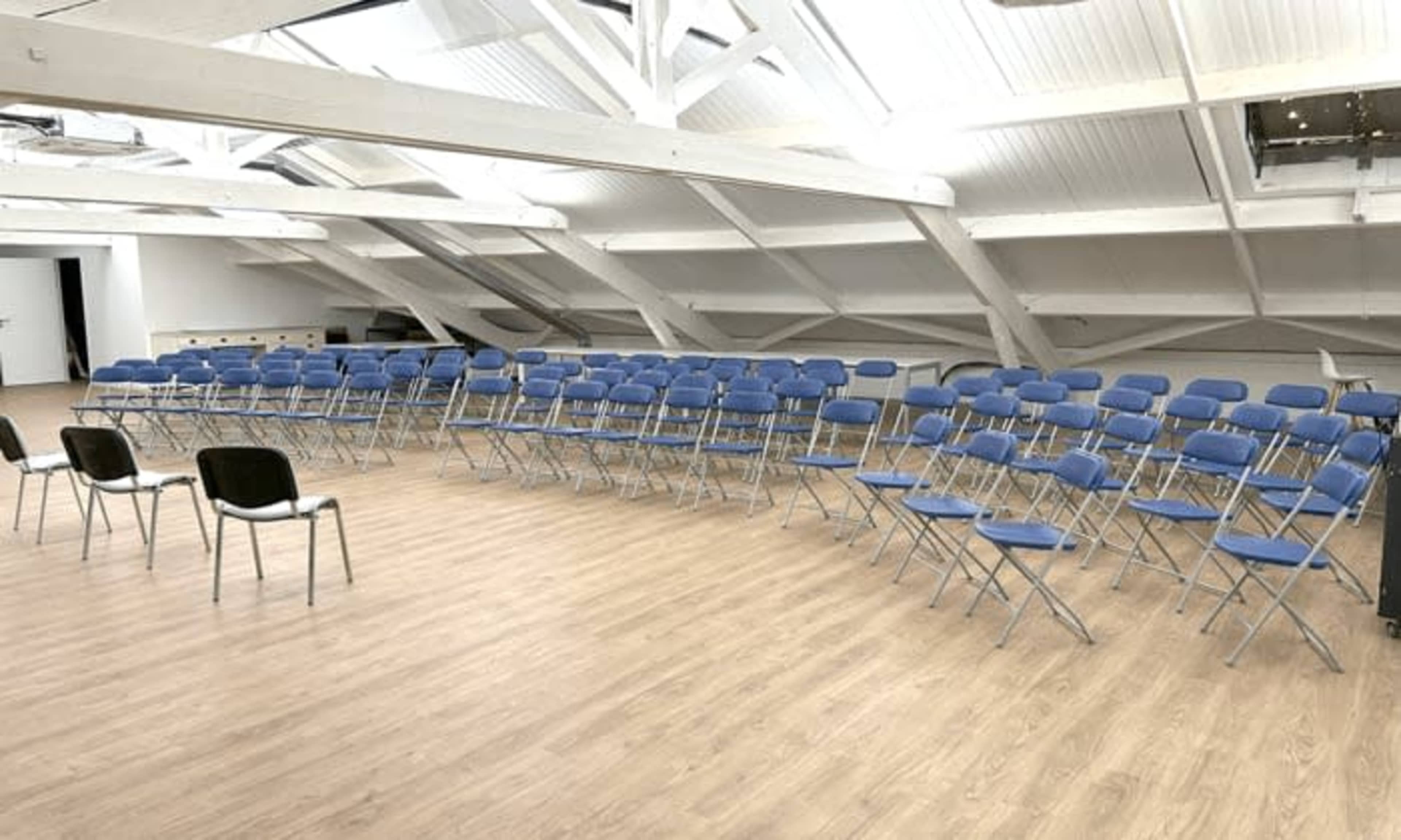 The image shows a large, open room with rows of blue folding chairs arranged in a semi-circle on a wooden floor beneath a sloped ceiling with exposed beams.