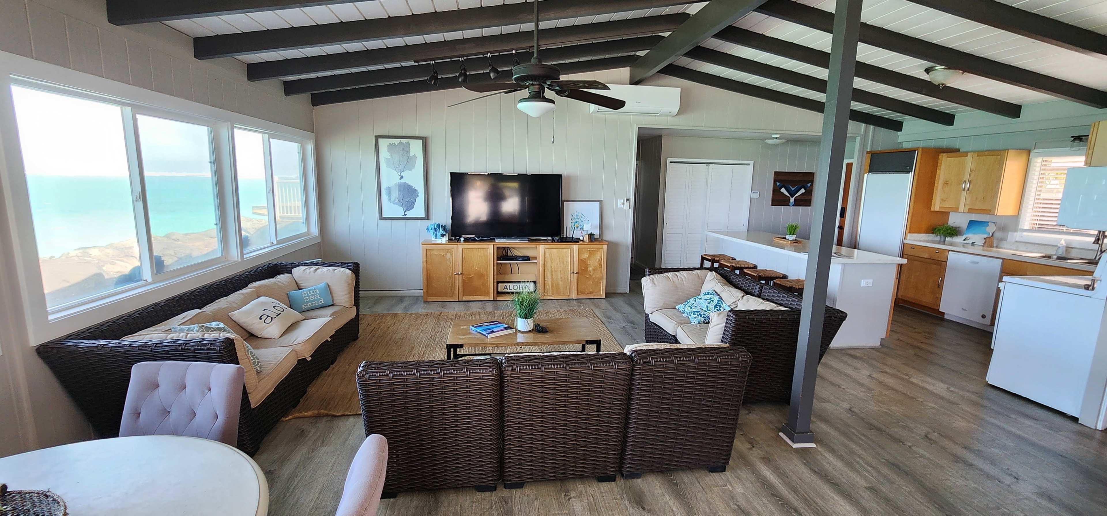 The image shows a coastal living space with two seating areas, a large television mounted on the wall, and an open kitchen visible in the background.
