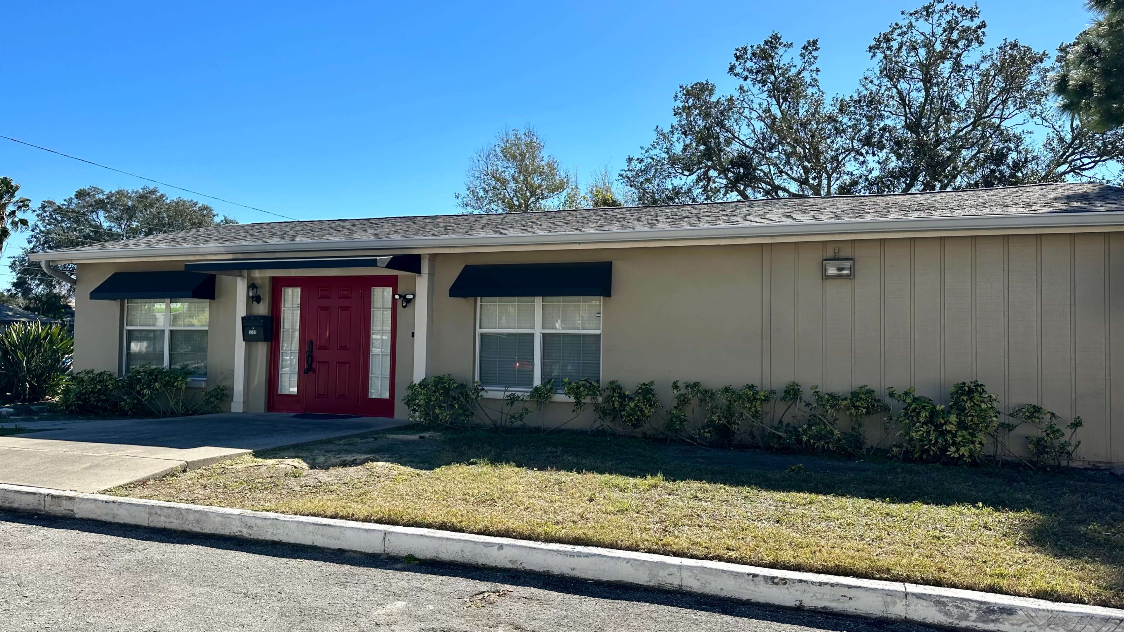 A single-story house features a red front door, black awnings over the windows, and a neatly maintained lawn.
