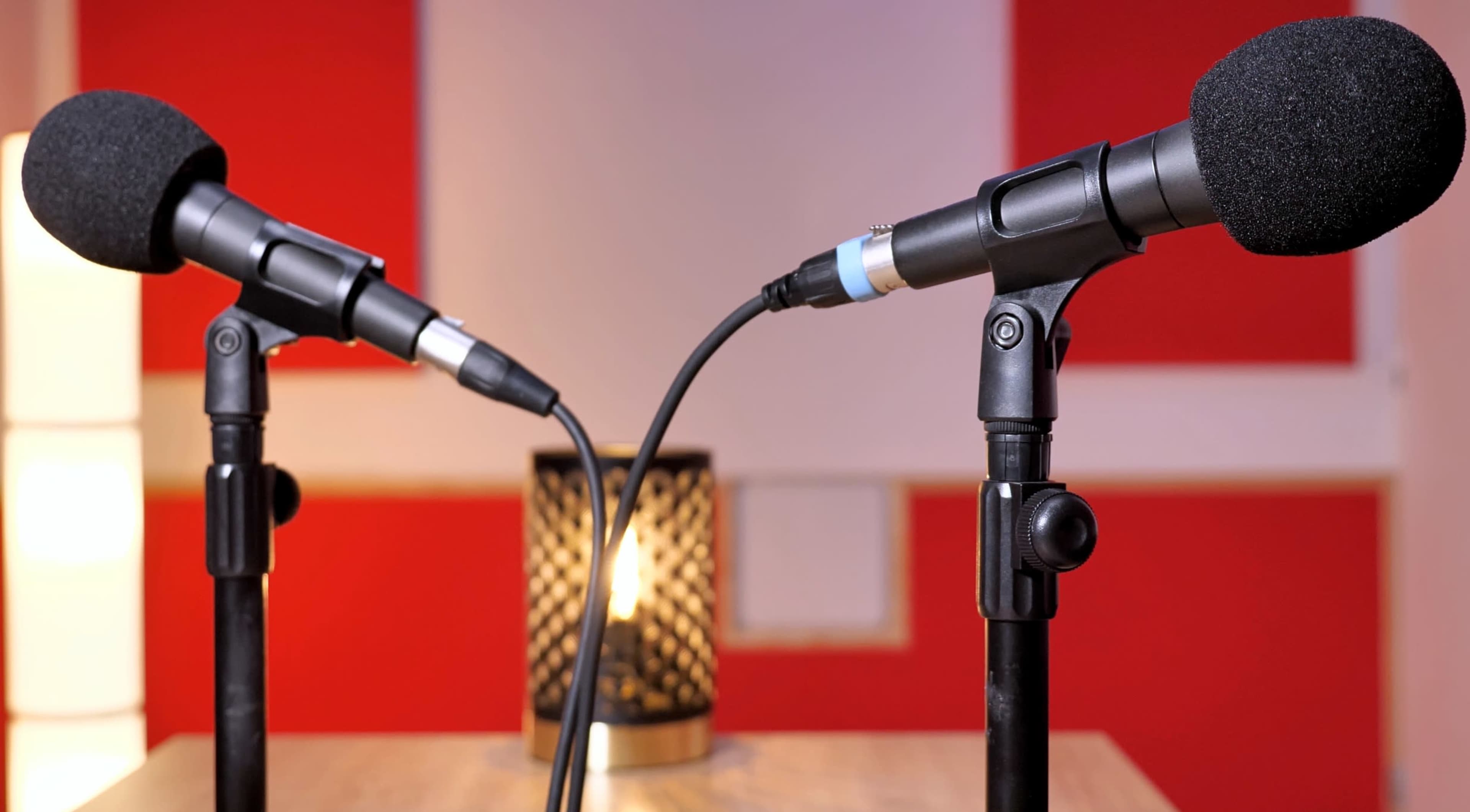 Two microphones on stands are positioned facing each other in front of a candle-lit lamp against a backdrop of red walls.