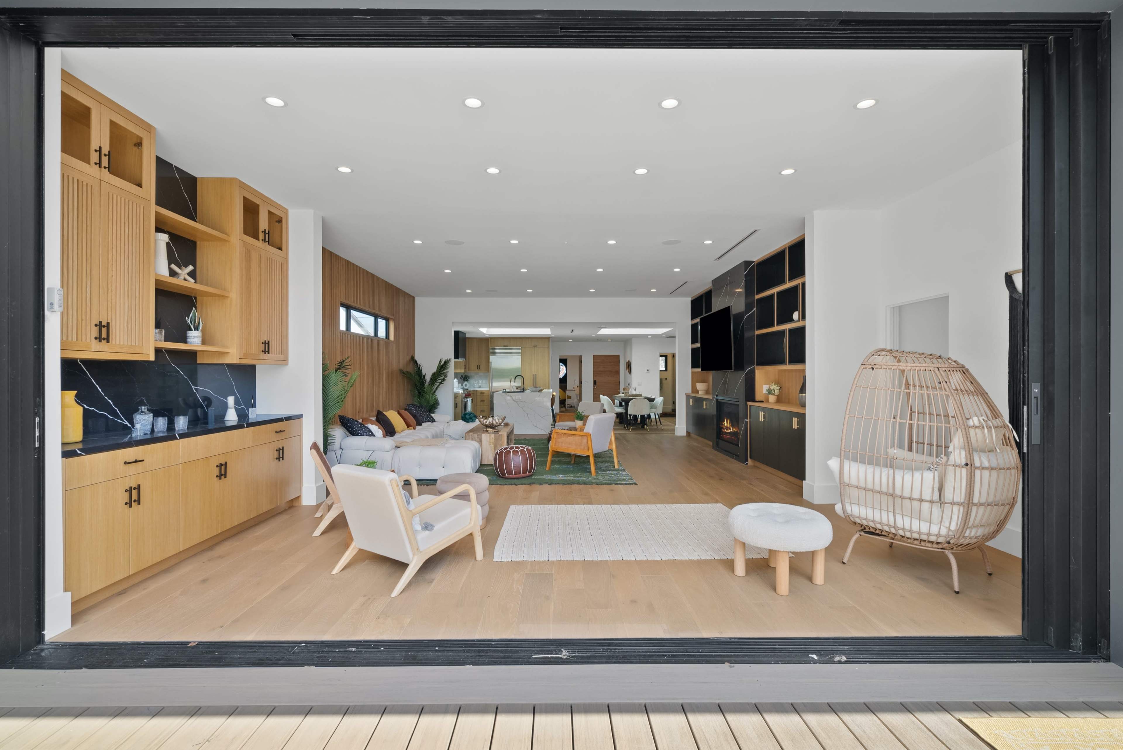 A spacious modern living room features large windows, wooden cabinetry, and a mix of contemporary furniture arranged around a central area rug.