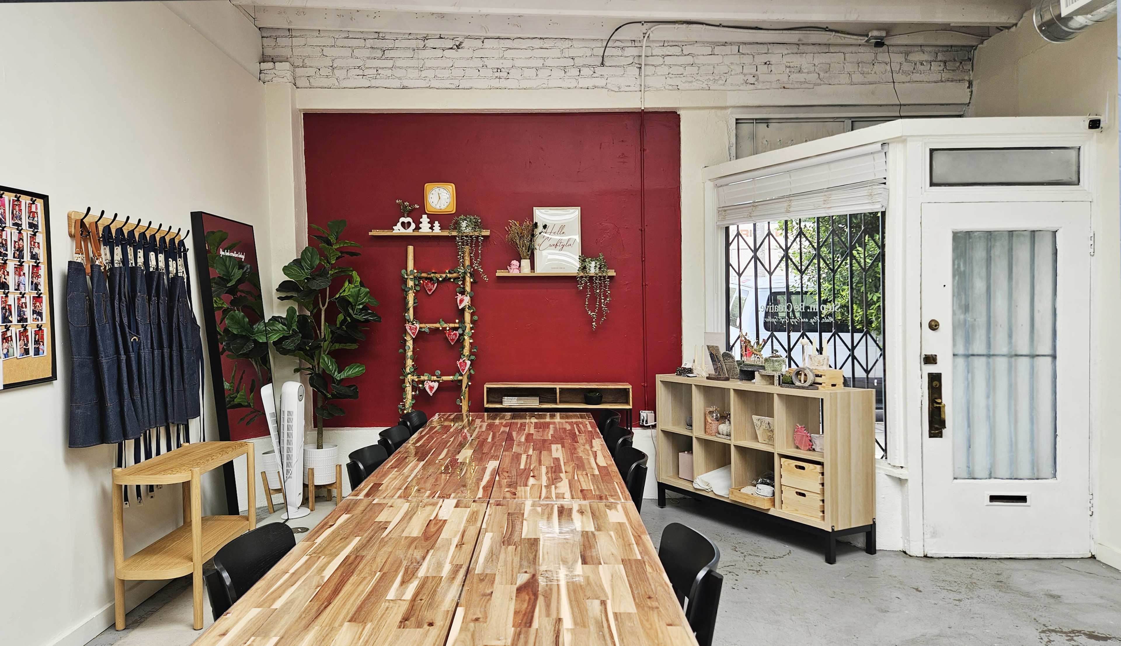 A spacious room features a long wooden table surrounded by black chairs, with a red accent wall, shelves holding decorative items, and a large window that opens to the outside.