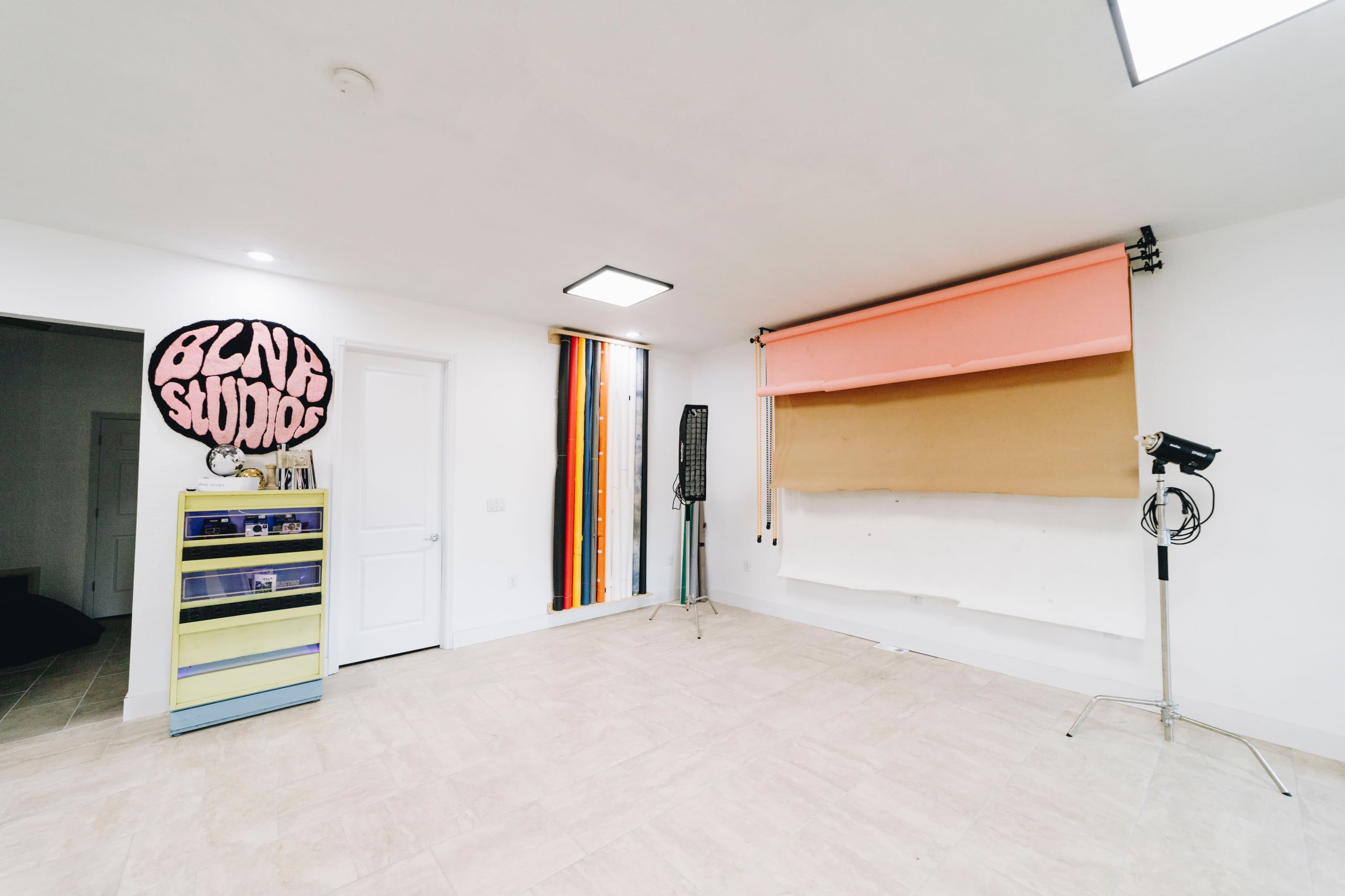 The image shows a spacious room with a white floor and walls, featuring a colorful backdrop, a tall light stand, and storage cabinets.