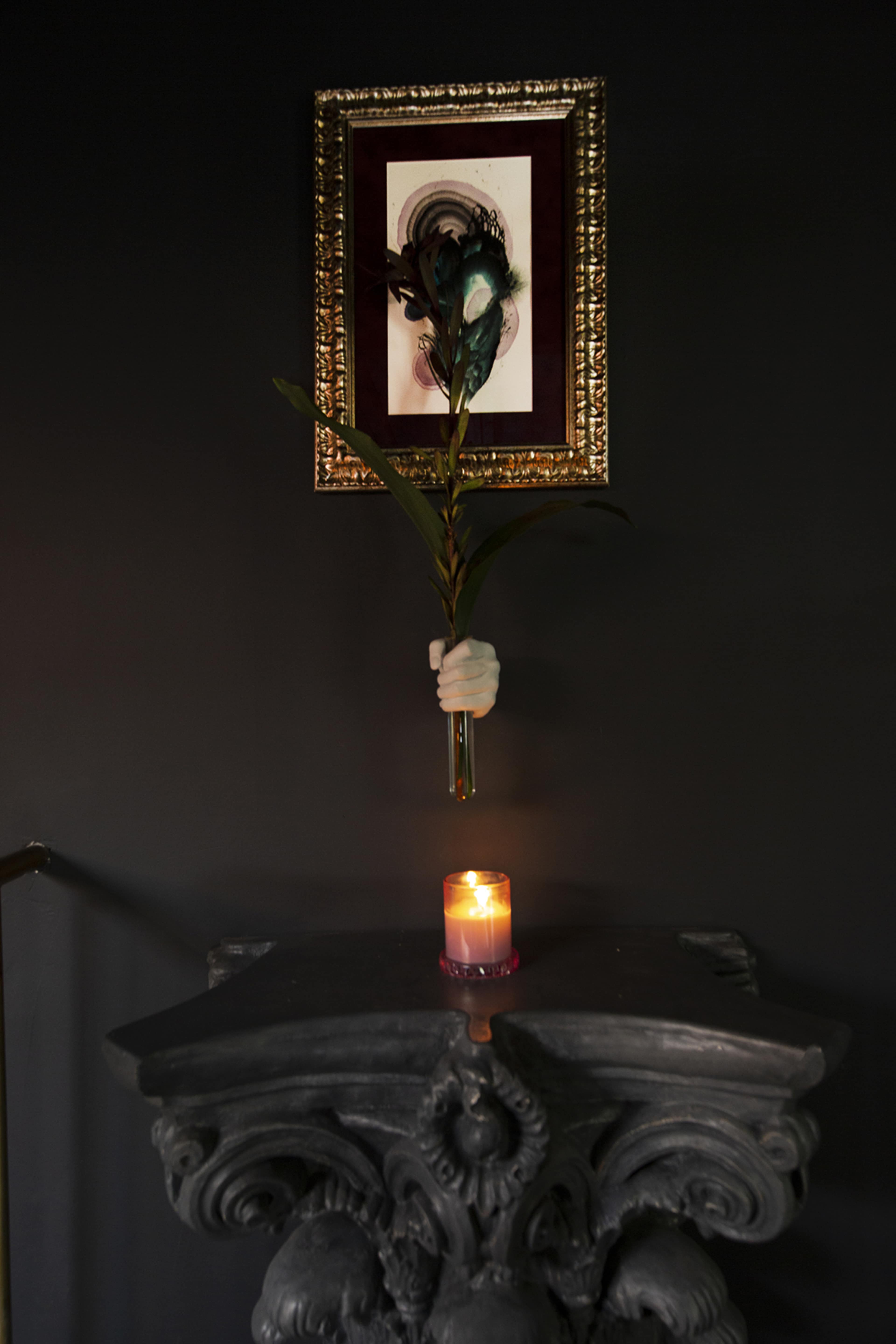 A hand-shaped vase holds a long-stemmed plant above a candle on a dark stone pedestal, with an ornate framed artwork in the background.