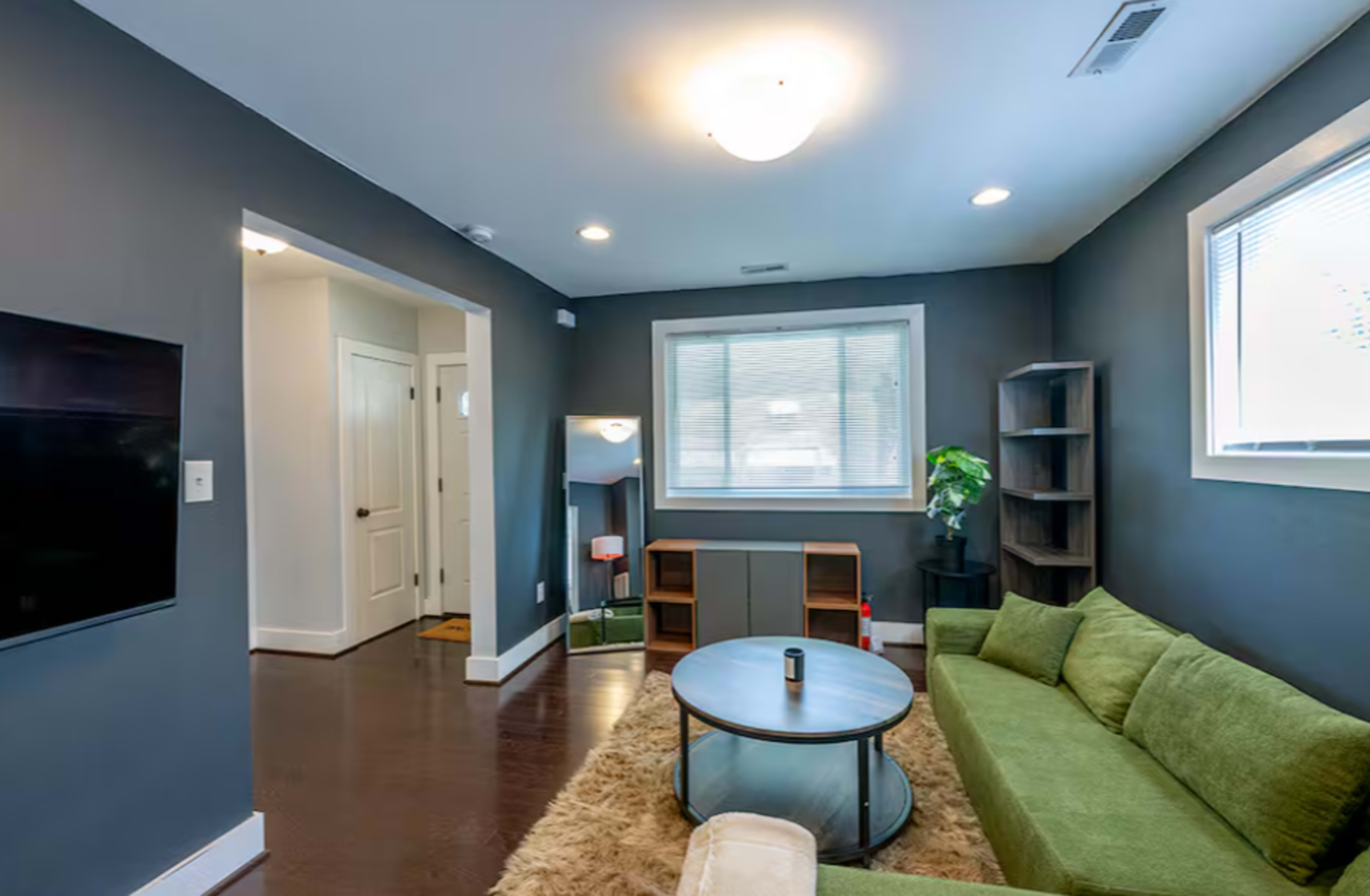 The image shows a modern living room with a green sectional couch, a round coffee table, and a large window letting in natural light.