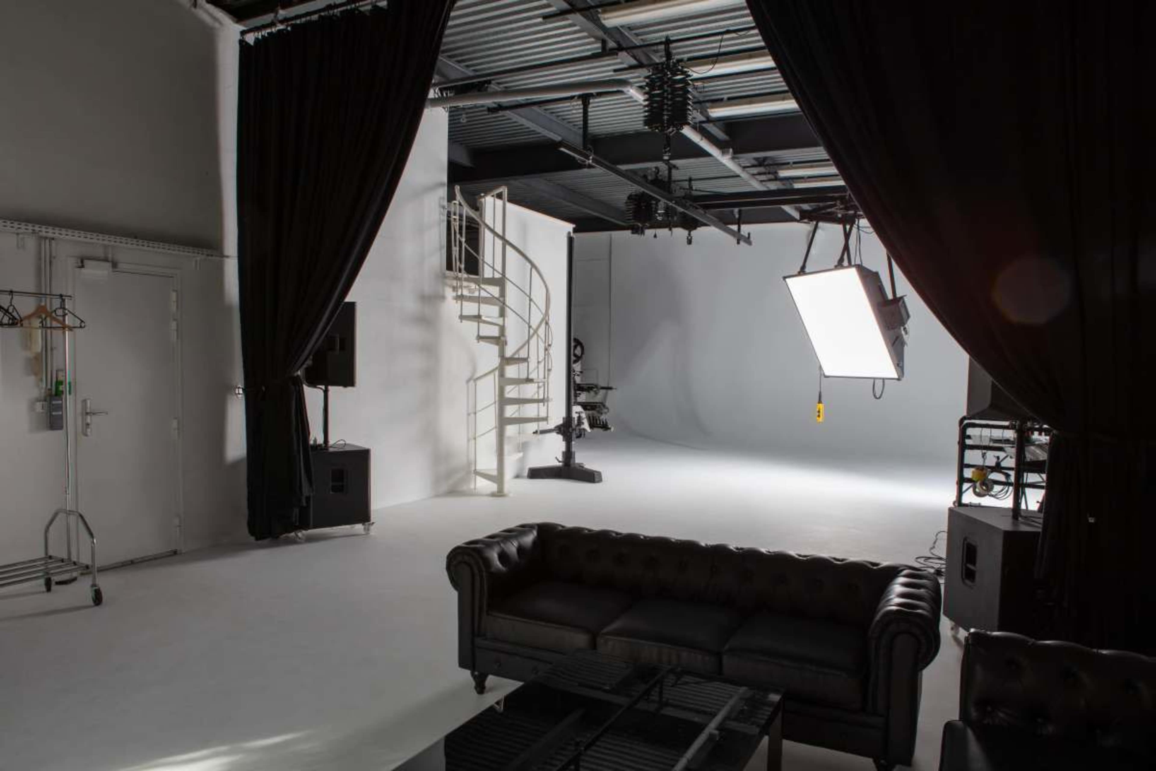 The image shows a spacious photography studio with a spiral staircase, lighting equipment, and a black leather sofa.