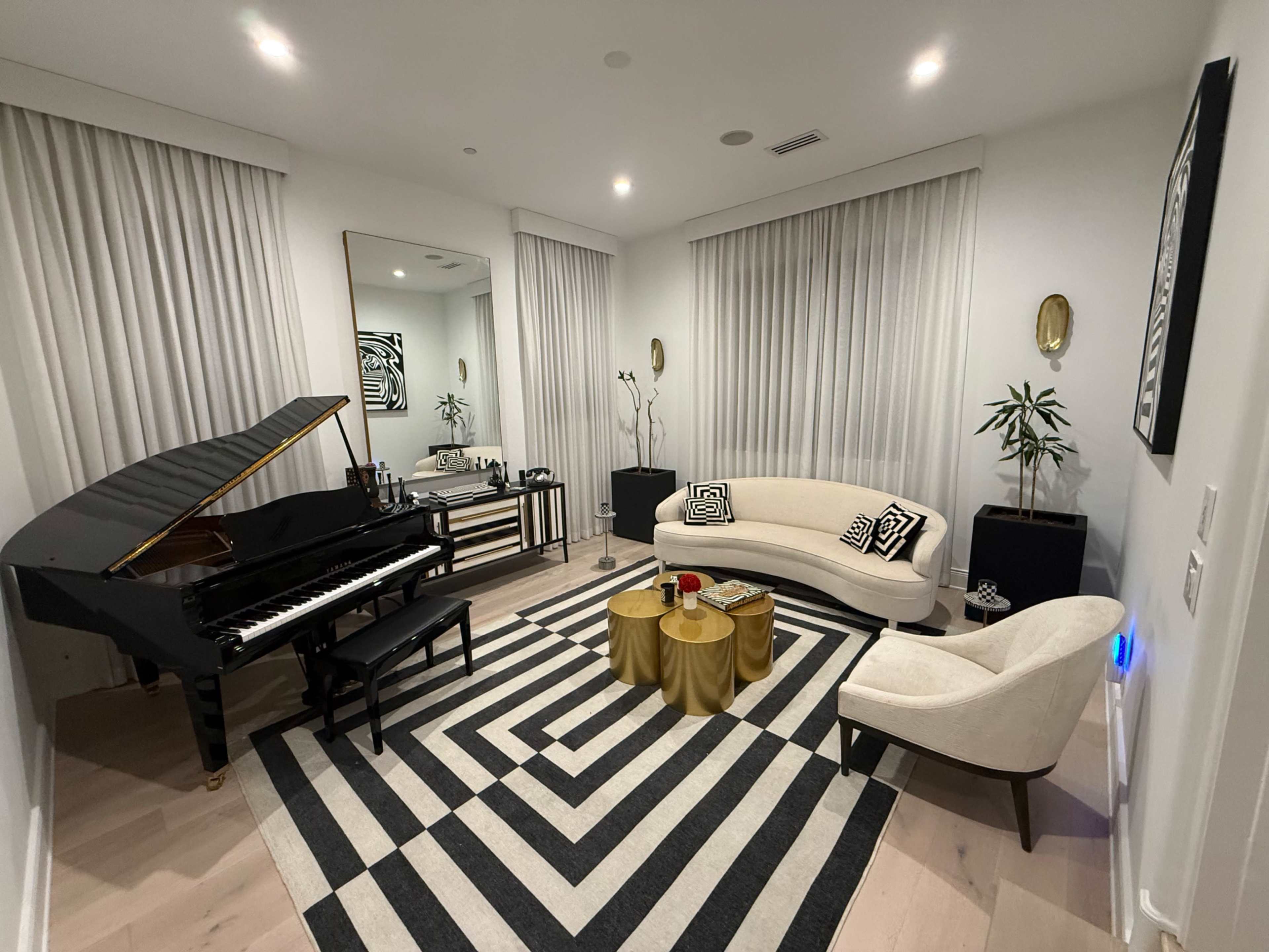 The image shows a modern living room featuring a grand piano, a curved sofa, two gold coffee tables, and large windows covered with sheer curtains.