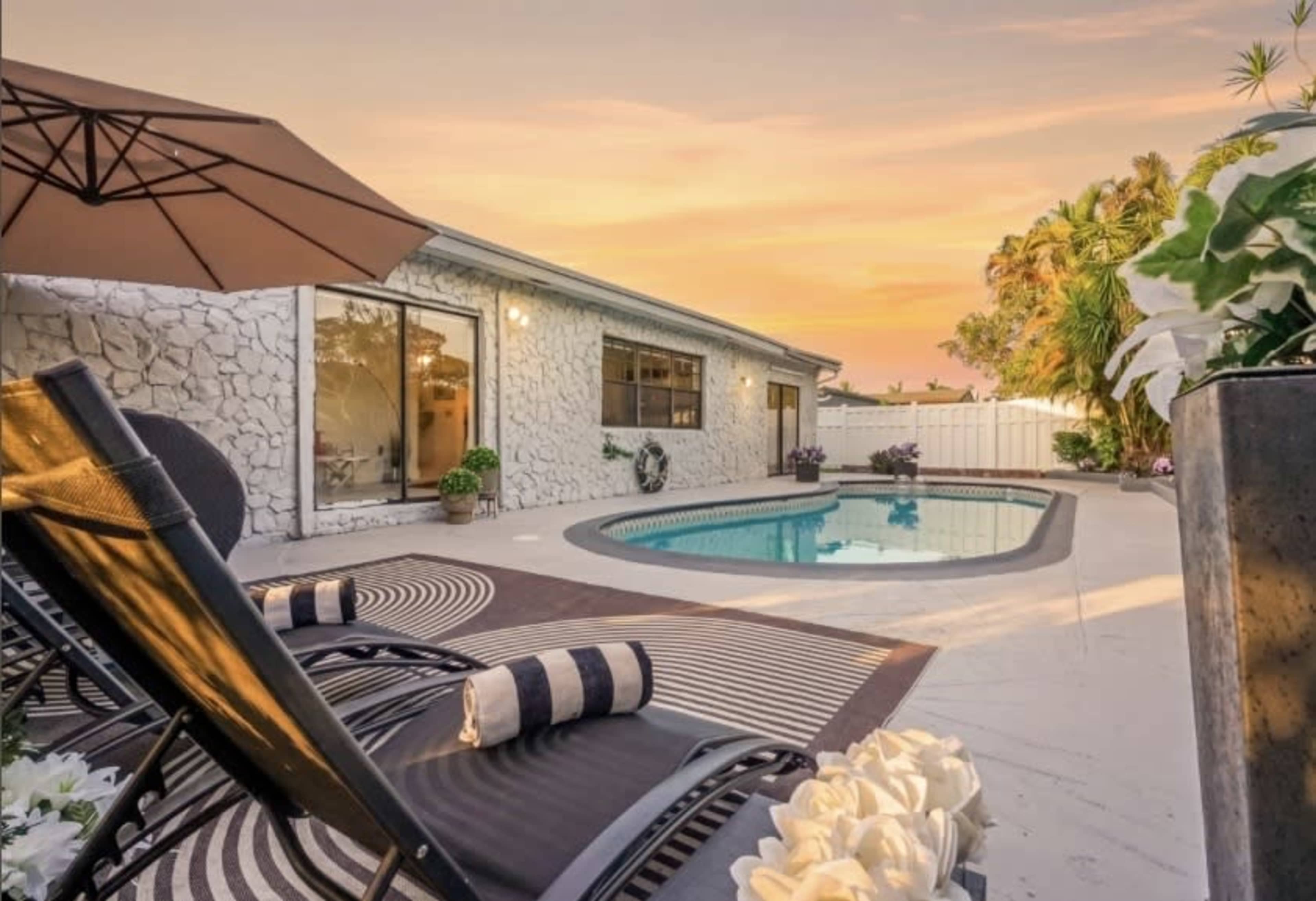 The image shows a backyard pool area with lounge chairs, an umbrella, and a house in the background during sunset.