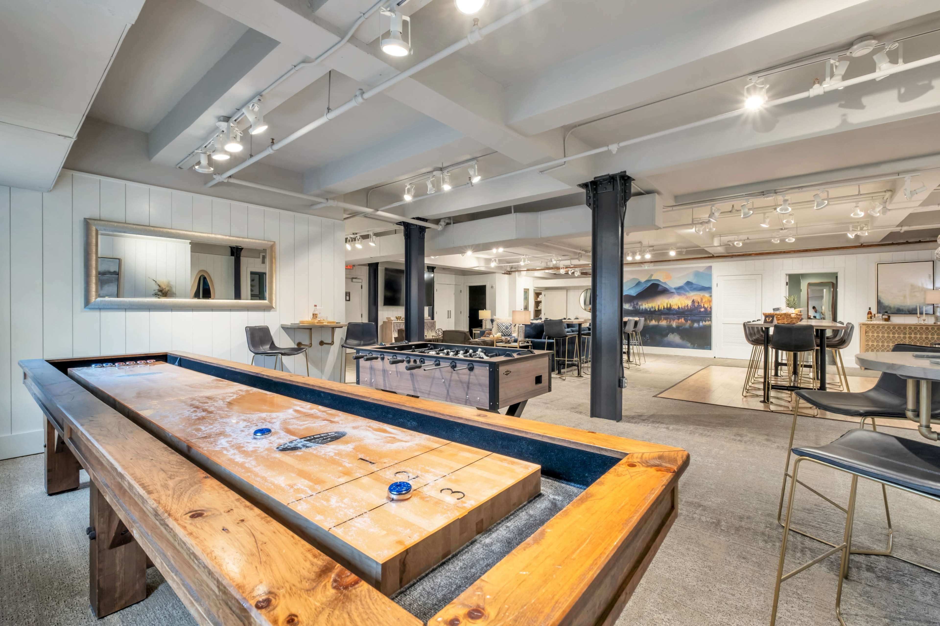 The image shows a modern recreational room featuring a shuffleboard table and a foosball table, with a bar area and art on the walls.