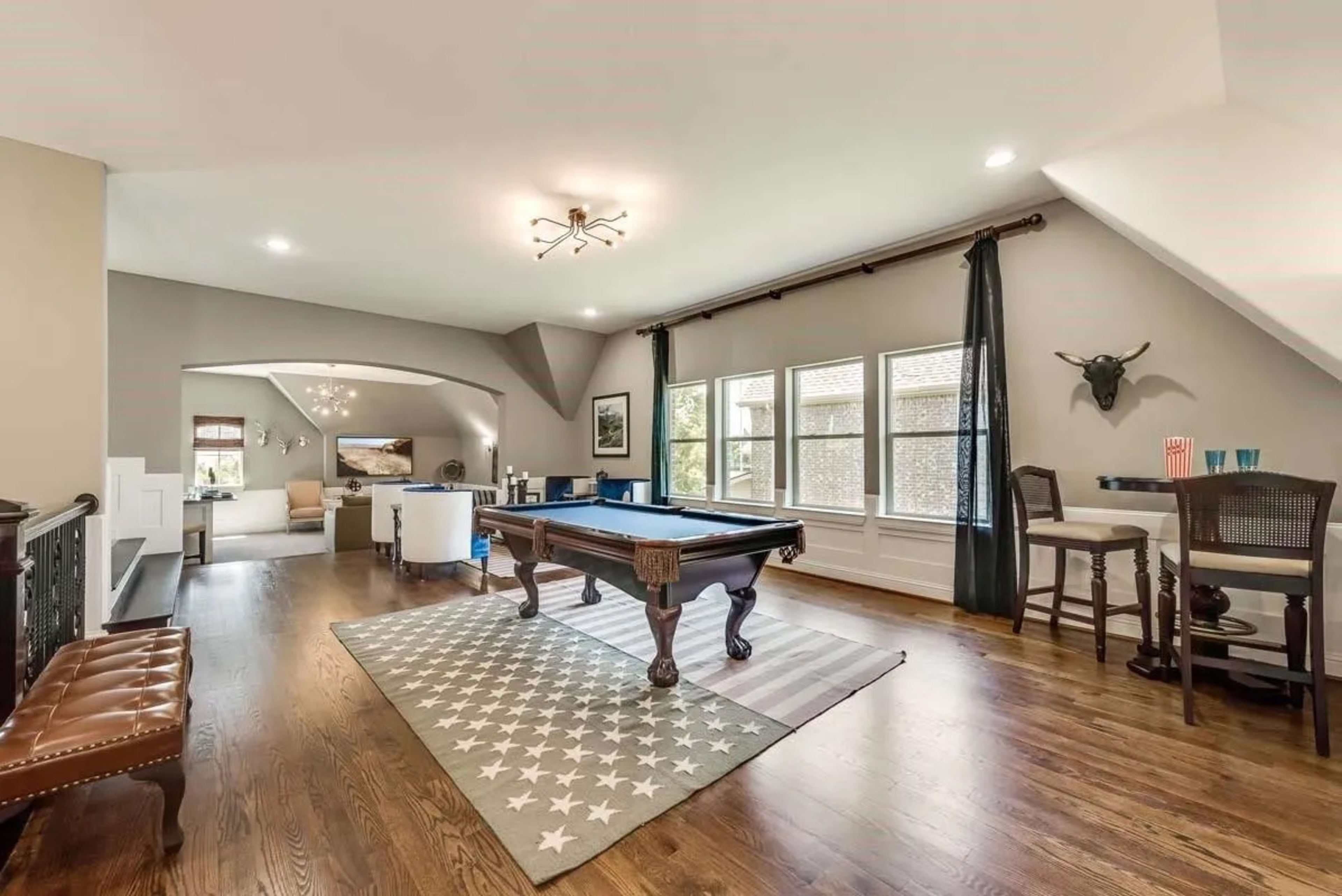 The image shows a spacious living area featuring a billiards table, a seating area, and large windows with natural light.