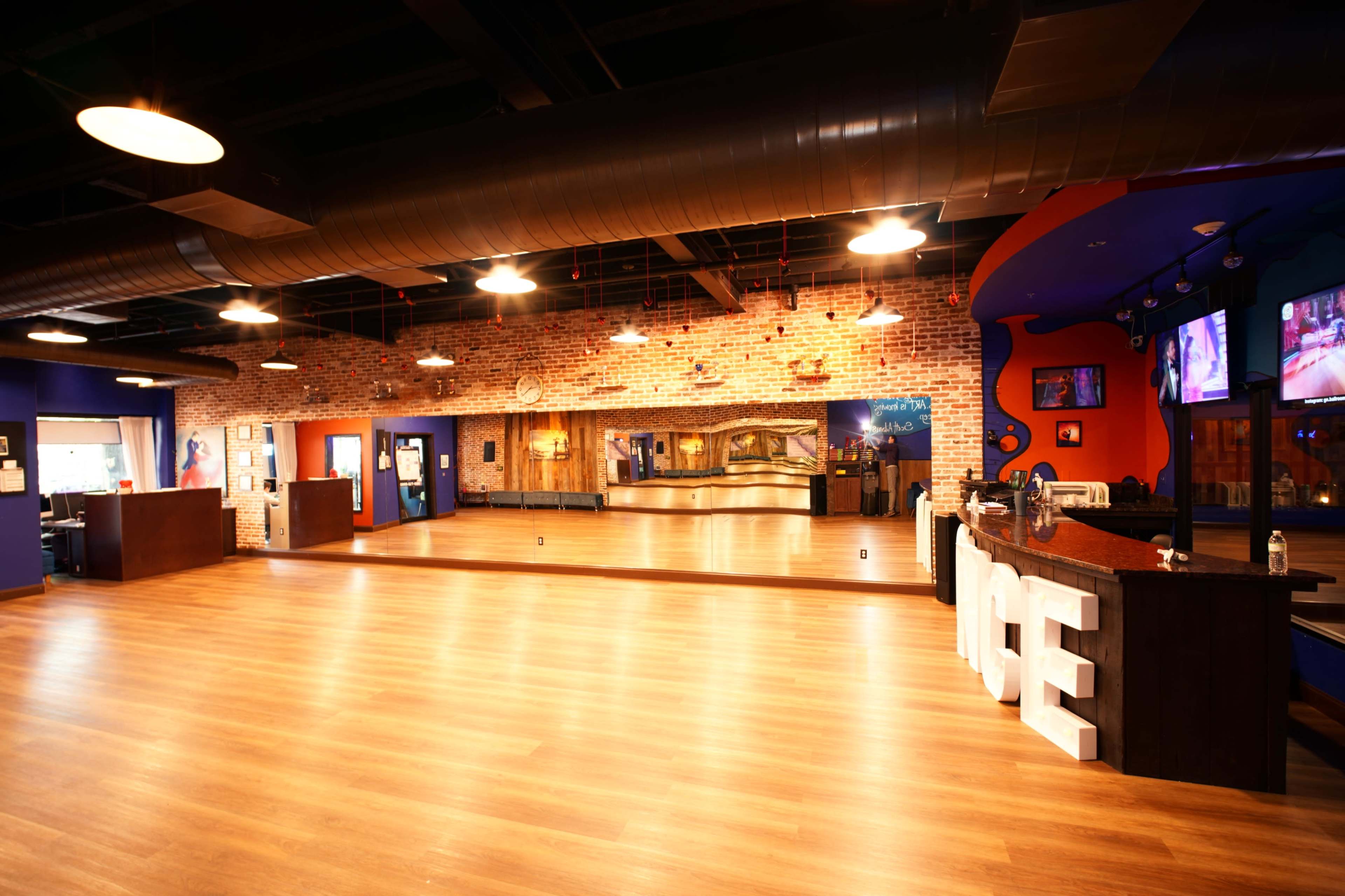 The image shows a spacious, brightly lit dance studio with wooden flooring, exposed brick walls, and a reception area featuring large letters that spell "DANCE."