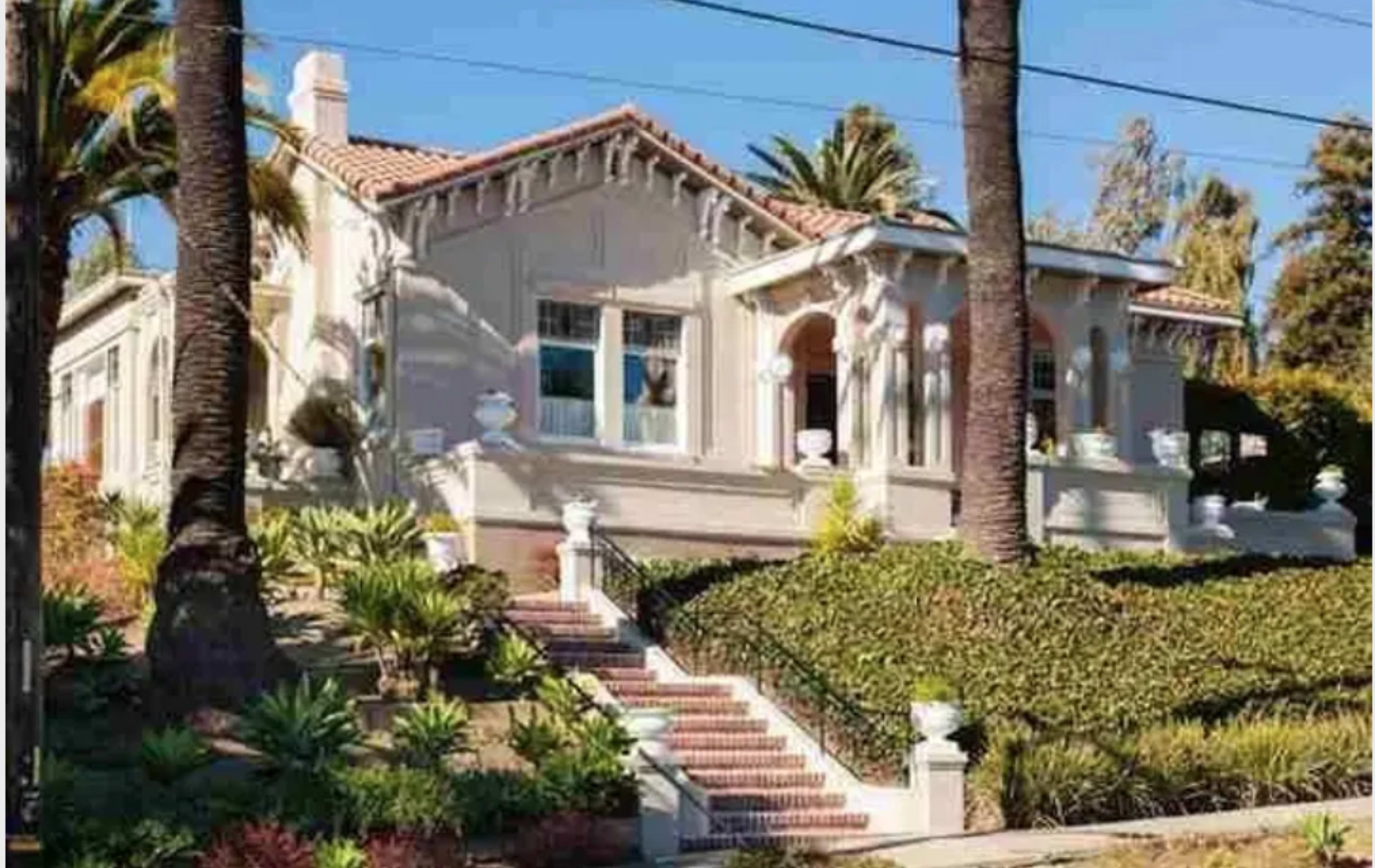 The image shows a large, decorative house with a tiled roof, surrounded by palm trees and well-maintained landscaping.