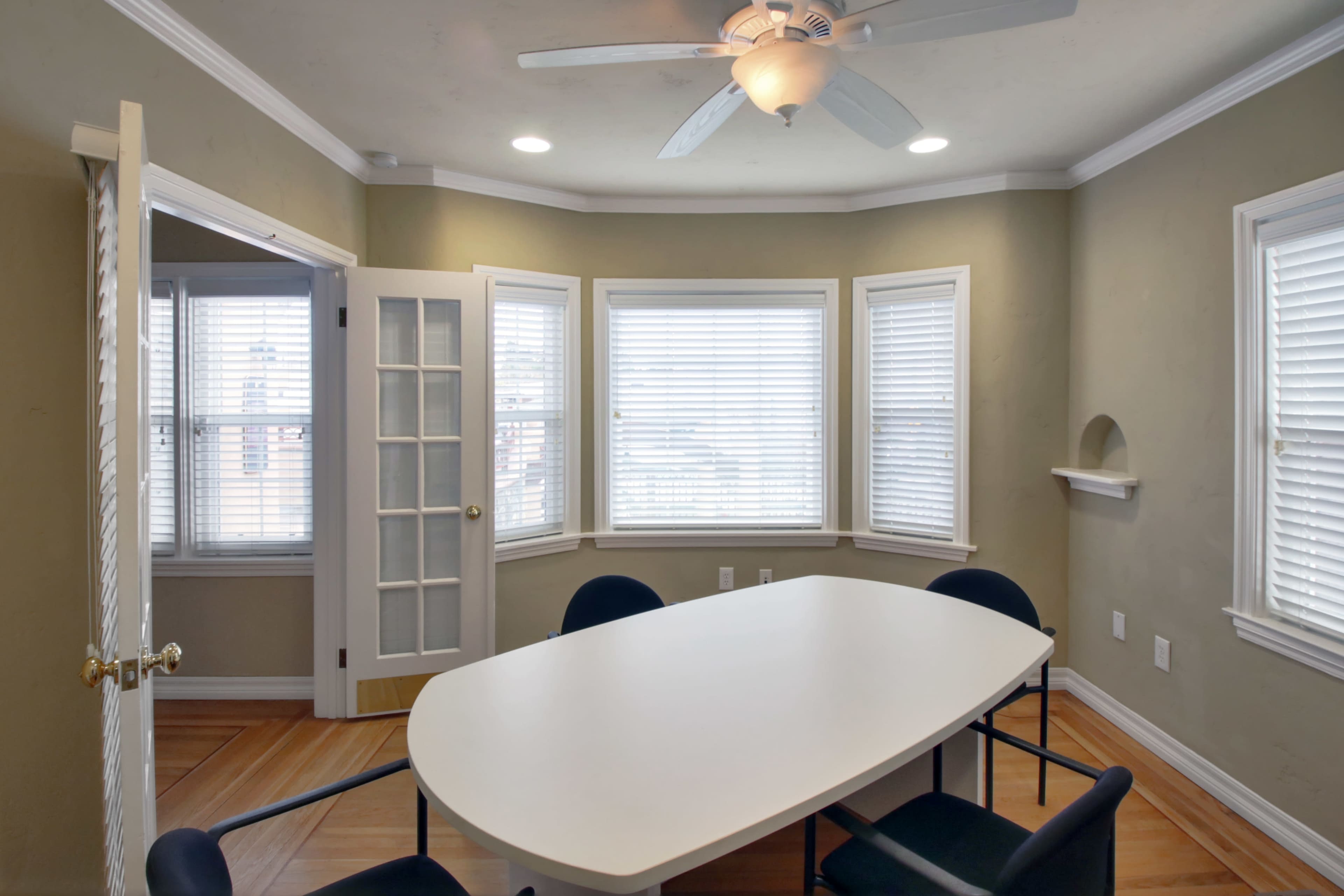 A bright room features a large white table surrounded by black chairs, with multiple windows allowing natural light to enter.
