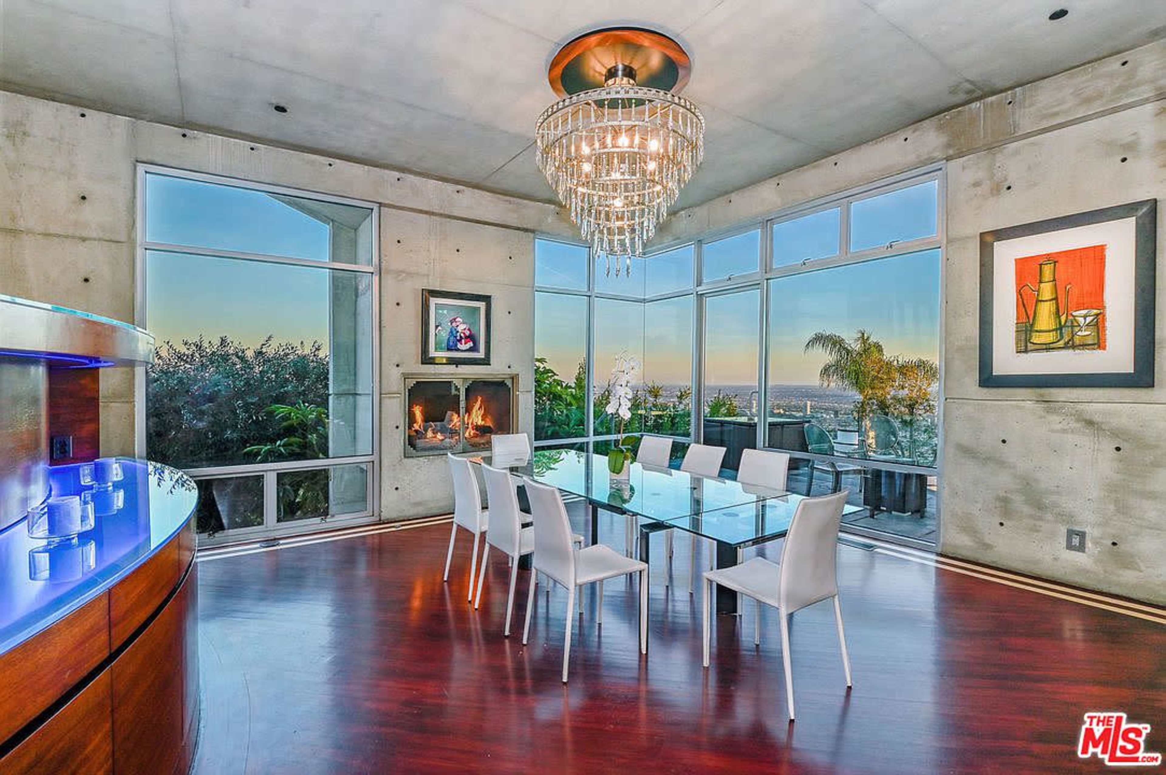 A modern dining room features a glass table surrounded by white chairs, with a large chandelier above and expansive windows offering a view of the outdoors.