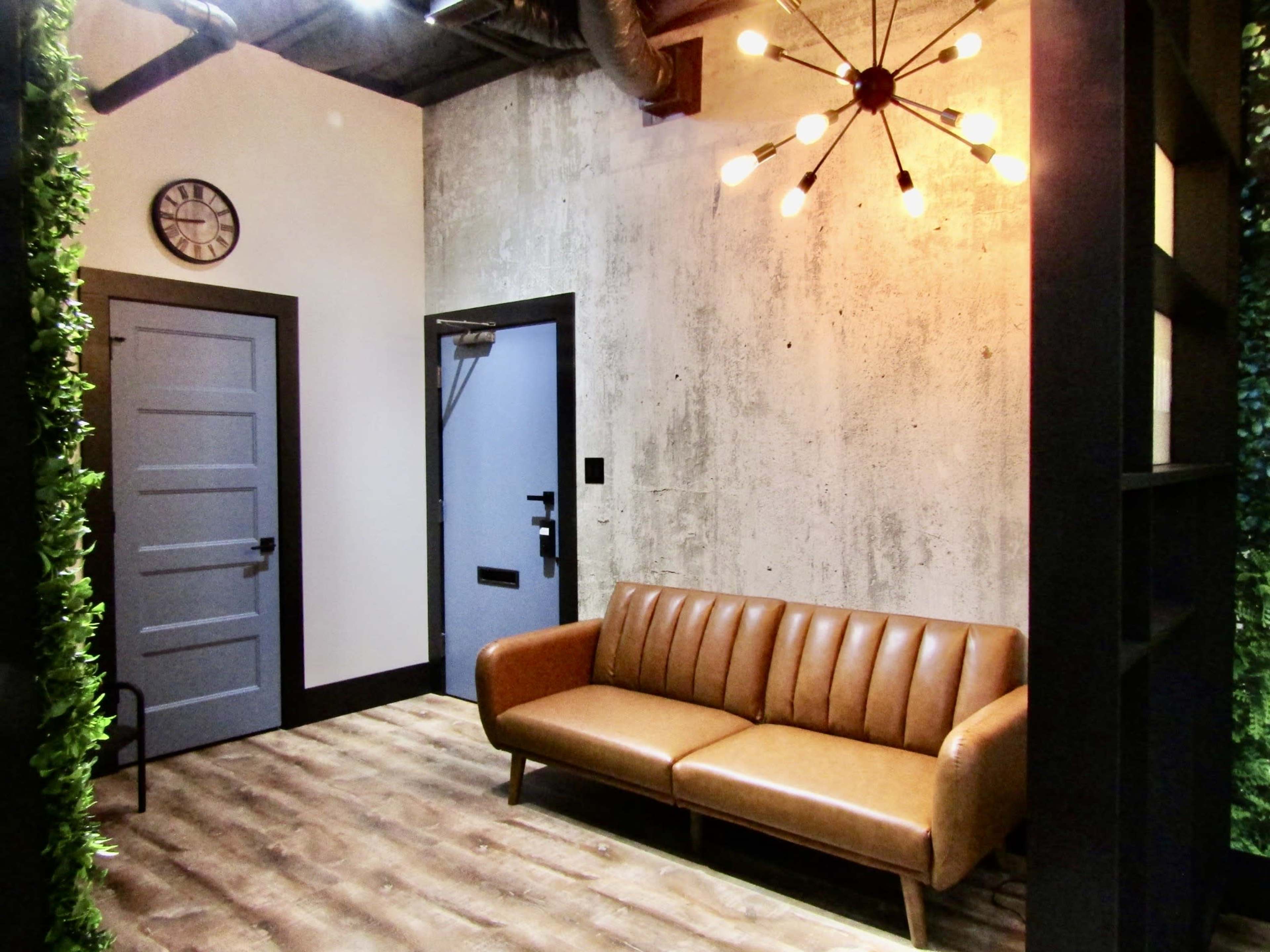 The image shows a modern interior space with a brown leather couch, a round wall clock, a blue door, and a concrete wall, surrounded by green foliage.