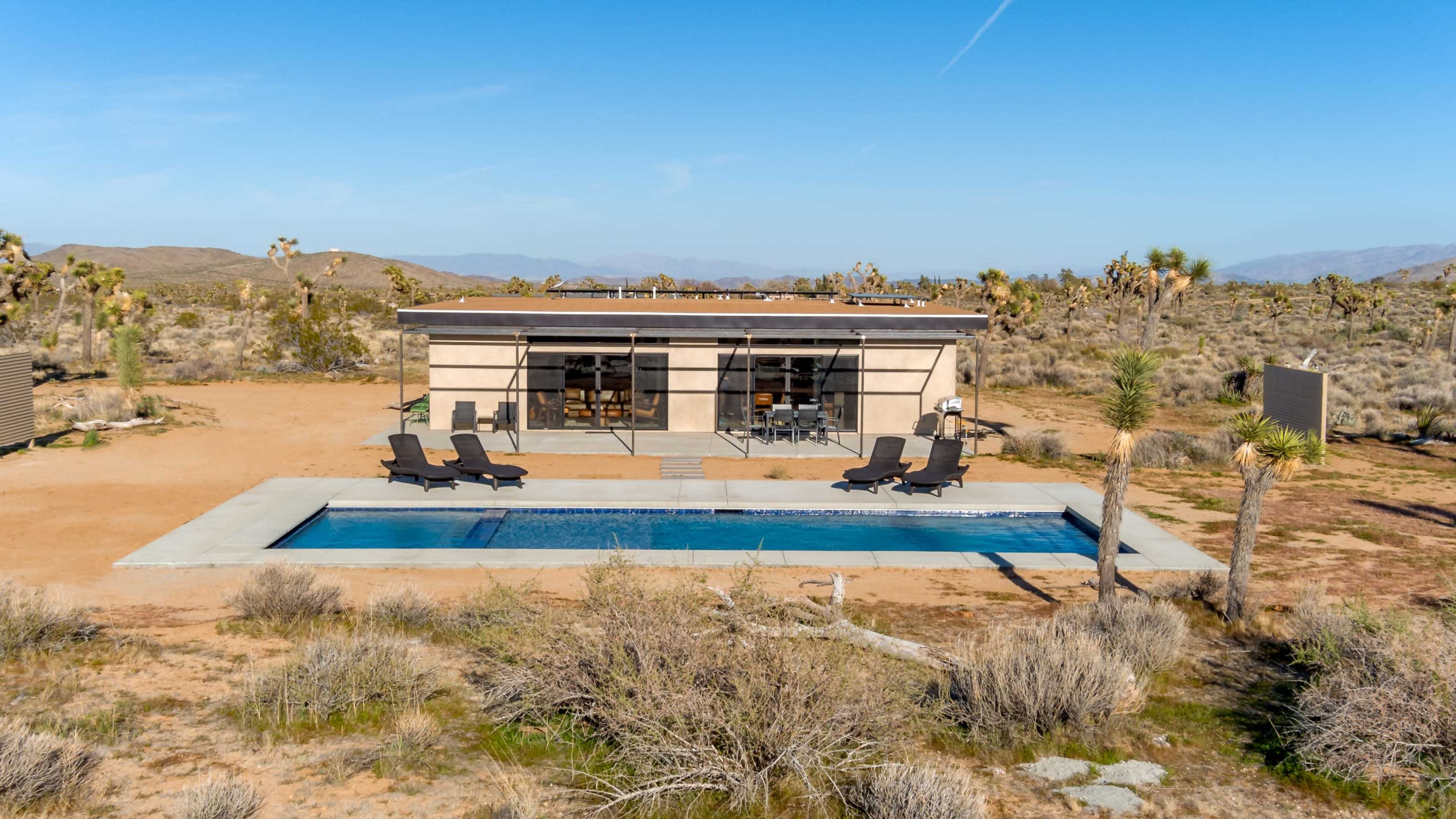 A modern single-story house with large windows overlooks a swimming pool in a desert landscape.